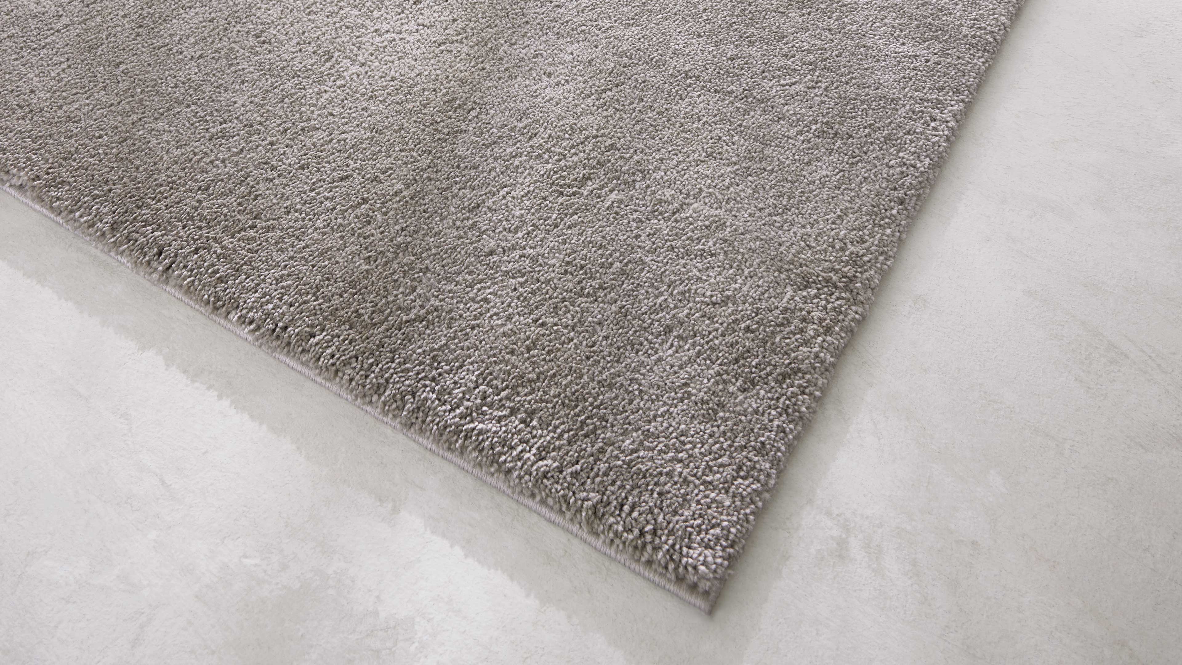 Eckansicht eines hellgrauen, dicht gewebten Teppichs mit kurzflorigem Flor auf hellem Betonboden.