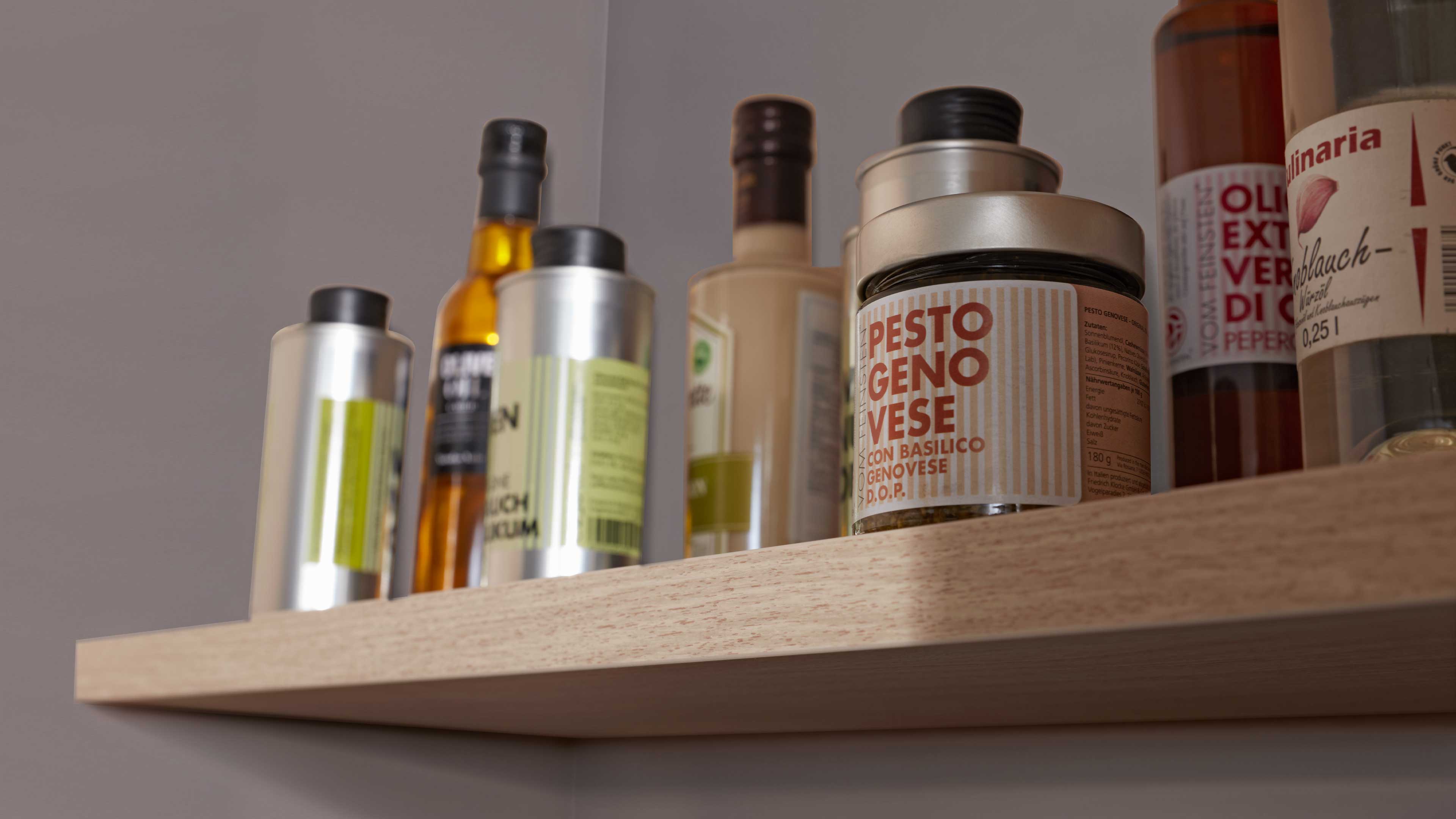 Holzregalbrett mit mehreren Glas- und Metallbehältern in verschiedenen Formen, darunter ein Glas mit Pesto-Etikett.
