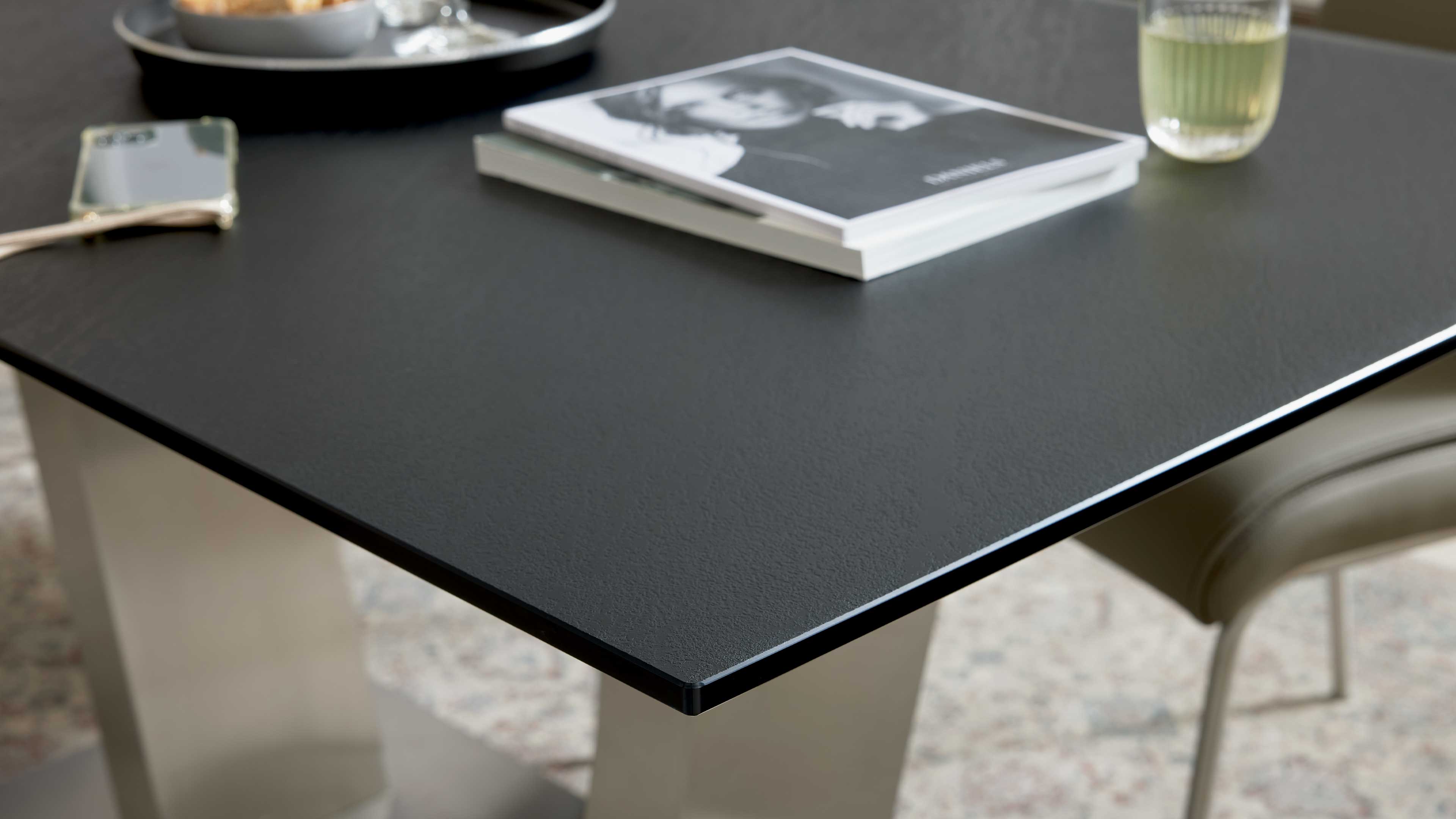 Schwarze, leicht strukturierte Tischplatte mit feiner Kante auf hellen Säulen, darauf zwei Bücher, Glas, Smartphone und ein Tablett.