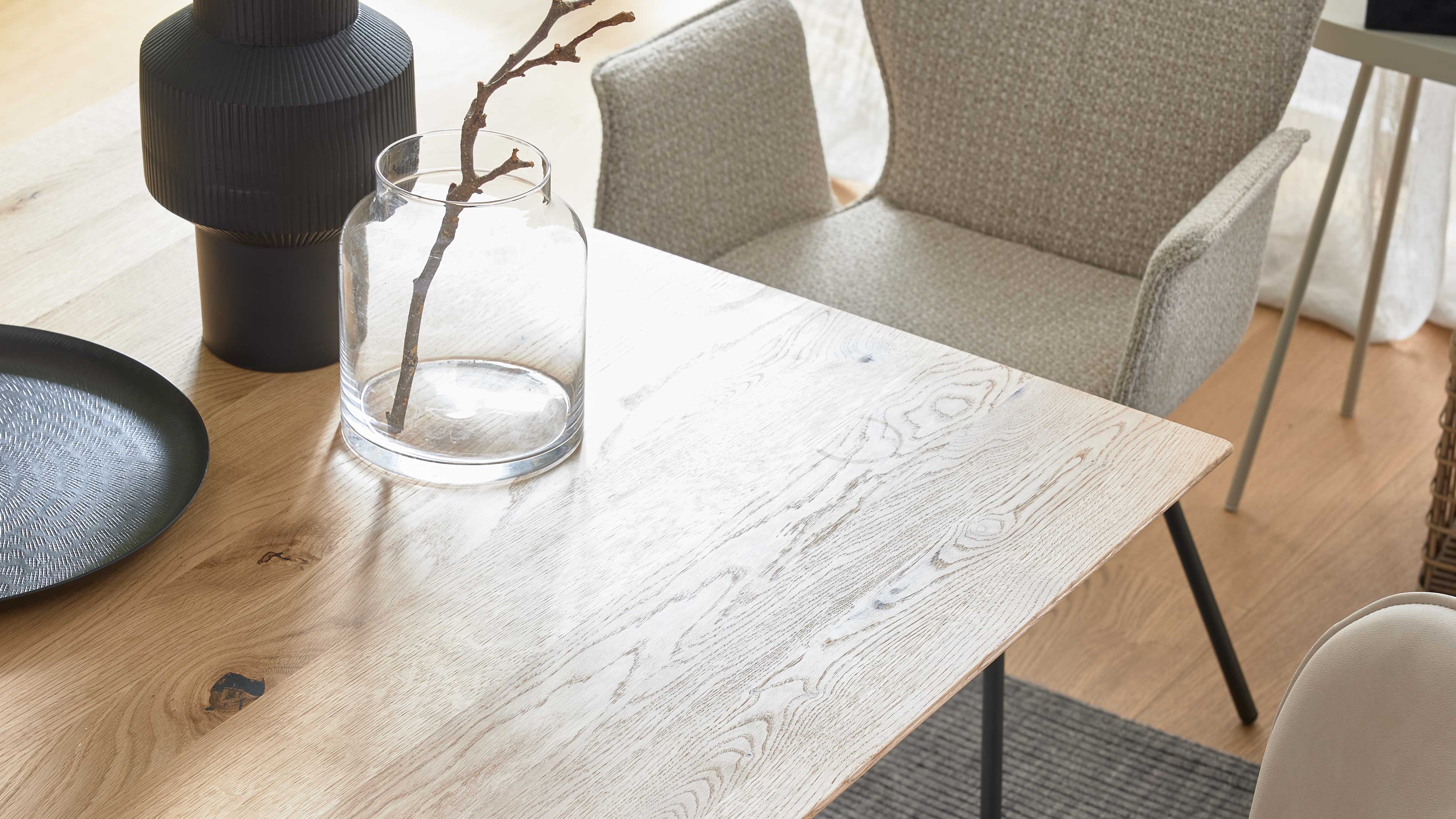 Heller Holztisch mit deutlicher Maserung, darauf eine gläserne Vase mit kahlem Zweig, eine schwarze Lampe und ein hellbeiger Sessel.