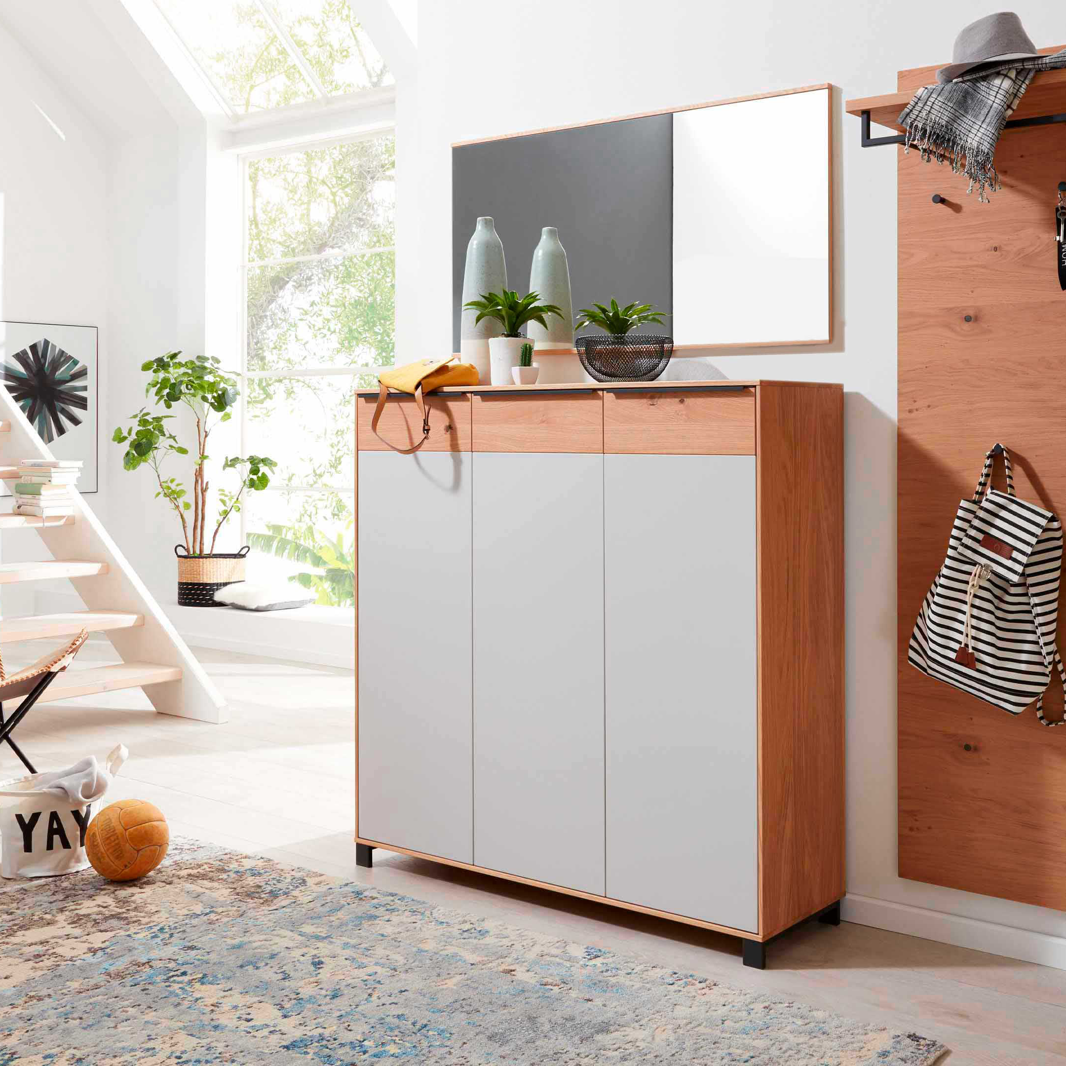 3-türiger Holzschrank mit grauen Fronten und Spiegel, darauf Vasen, Pflanzen; rechts Paneel mit Hut, Schal, Jacke und Rucksack.