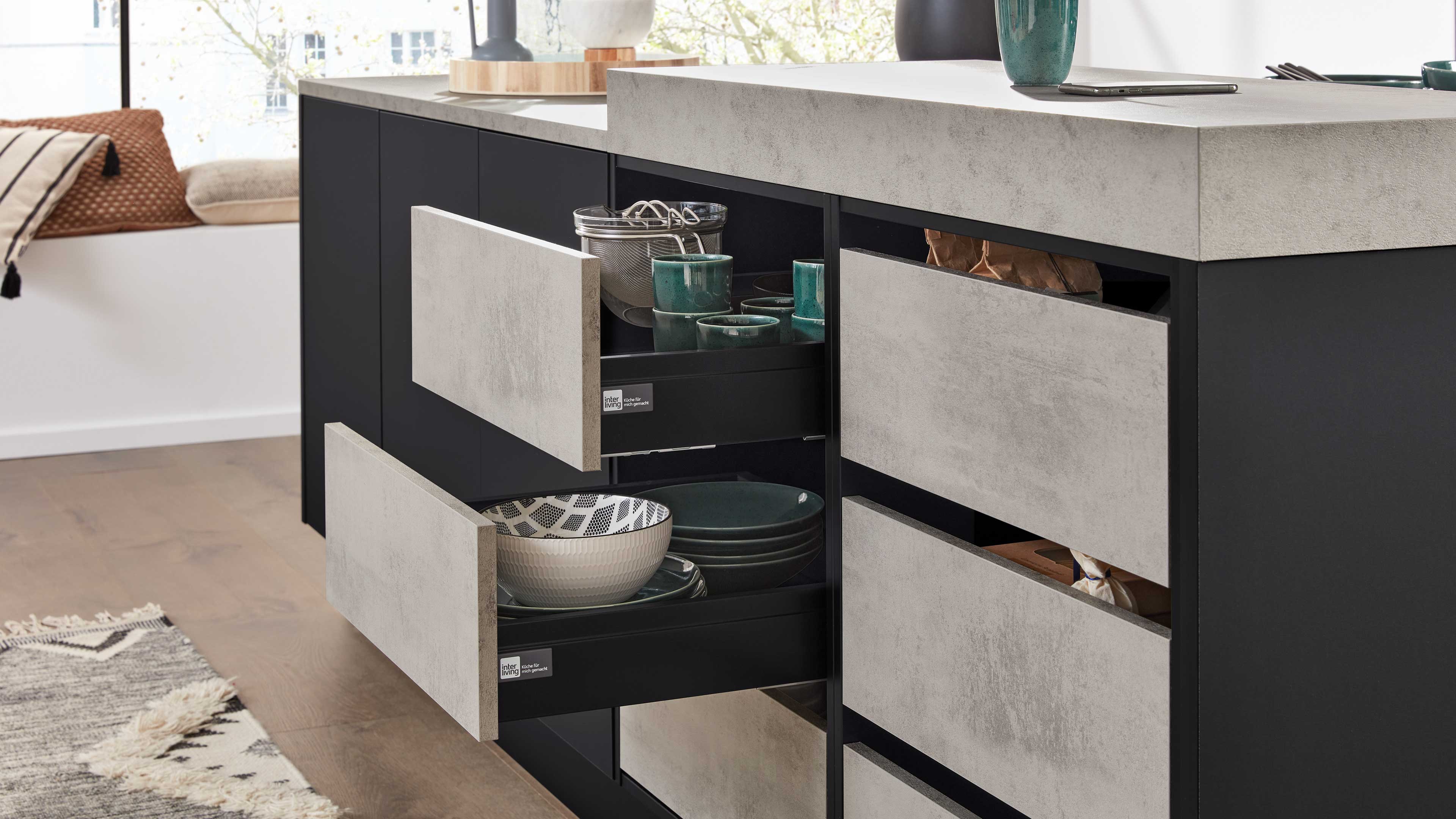 Offene Schubladen einer Kücheninsel mit Betonoptik-Fronten, gefüllt mit Schalen, Tellern und grünen Tassen, links Sitzbank mit Kissen.