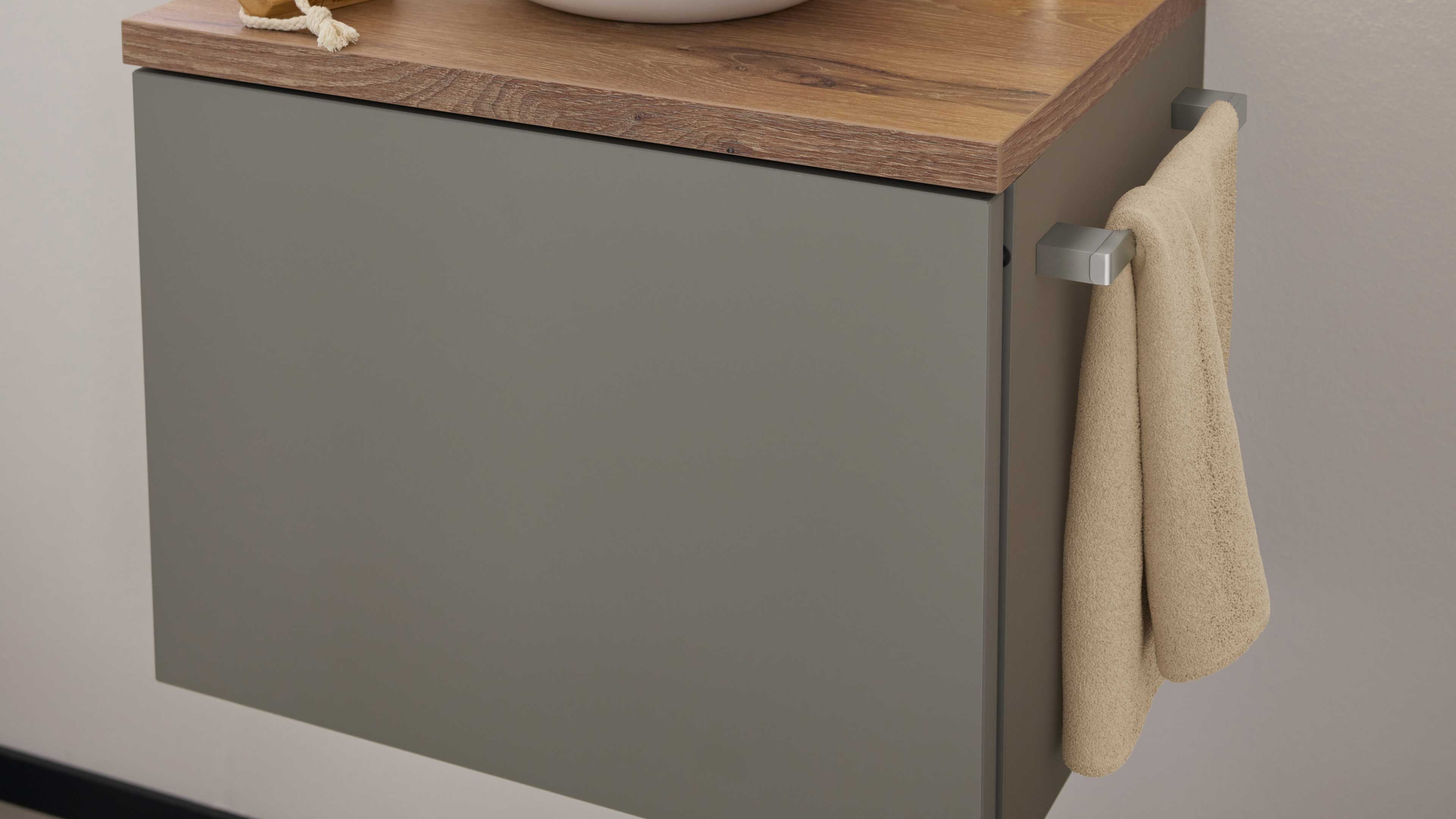 Hängender grauer Badunterschrank mit holzfarbener Arbeitsplatte und seitlichem quadratischem Metallhandtuchhalter mit beigem Handtuch.