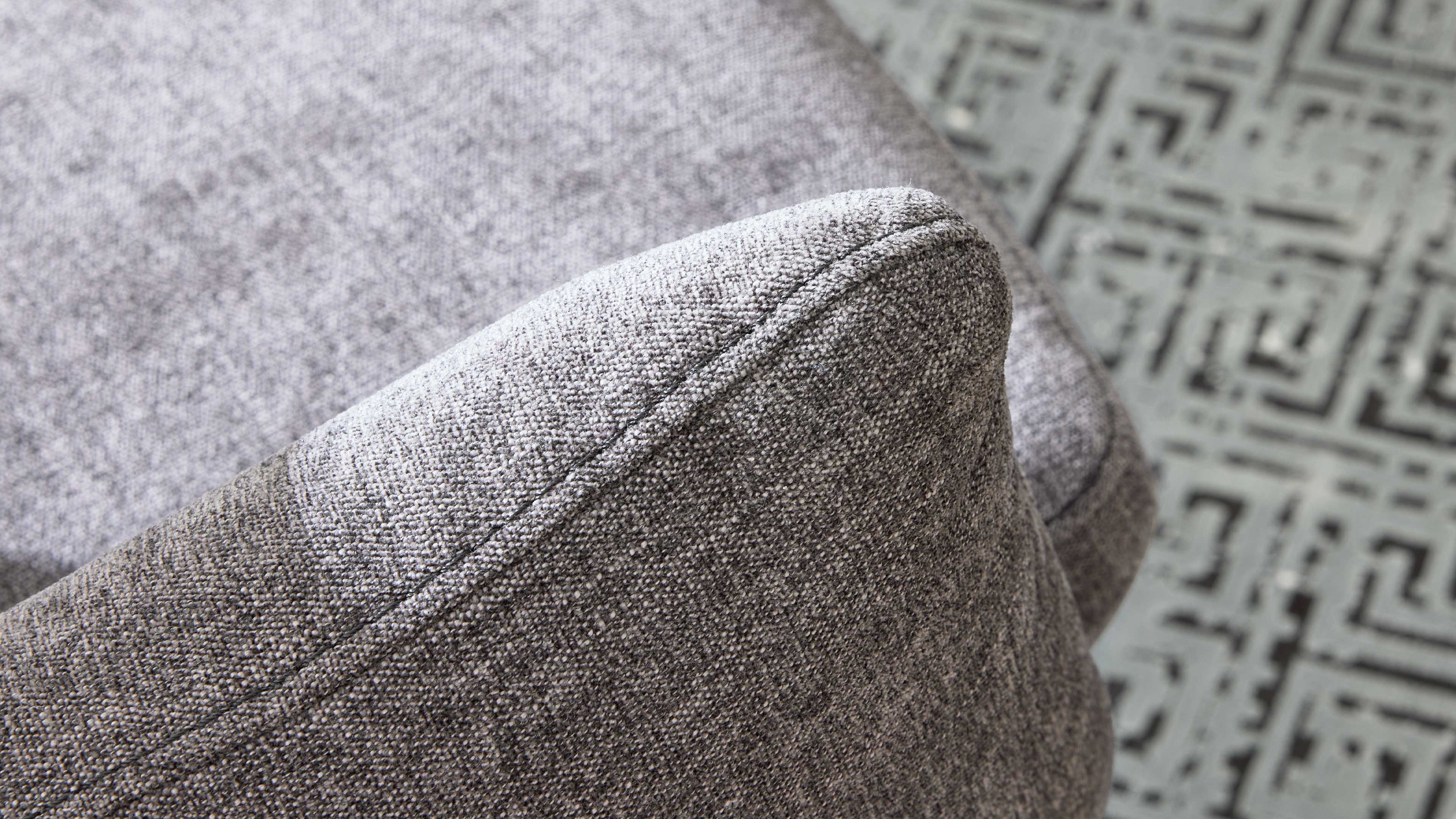 Nahaufnahme der abgerundeten Armlehne eines hellgrauen Sofas mit grob gewebtem Stoff und sichtbarer Naht, Musterteppich im Hintergrund.
