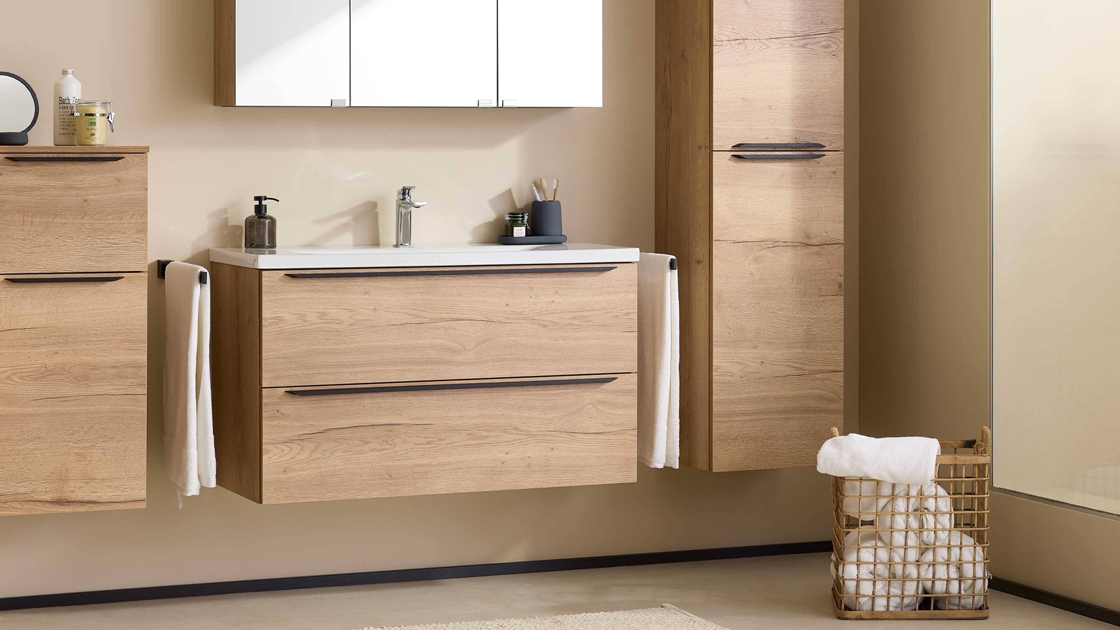 Hängender Waschtischunterschrank in Eicheoptik mit weißer Keramikplatte und Chromarmatur, rechts hoher Schrank und Drahtkorb.