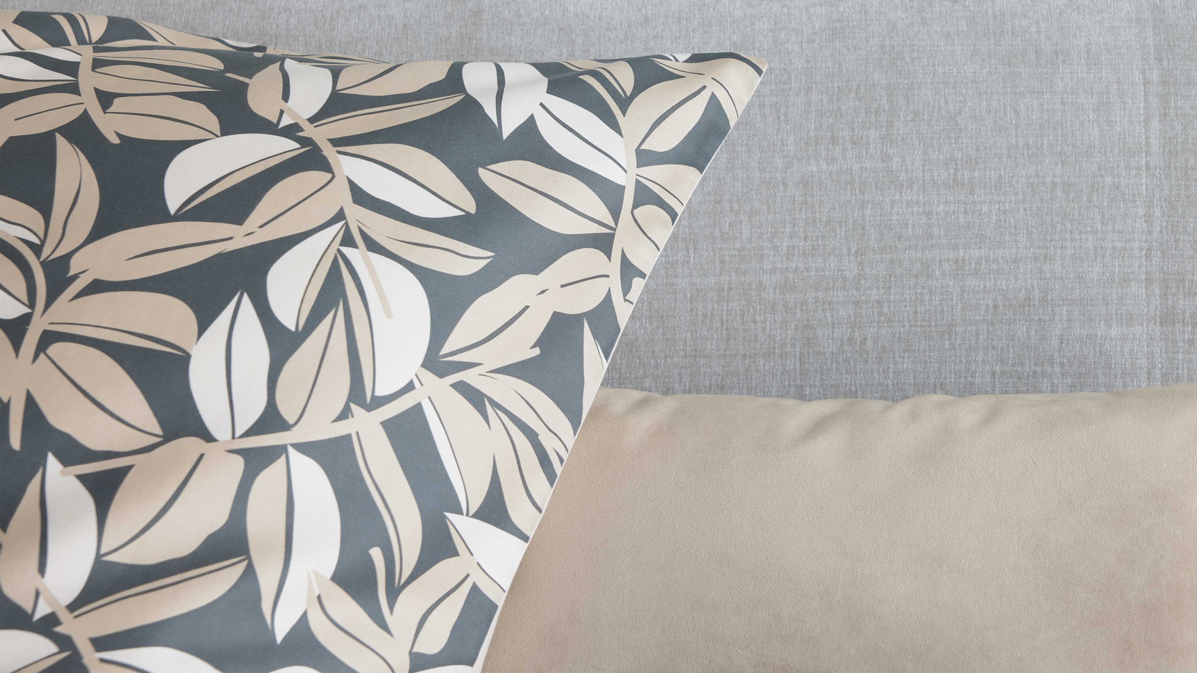 Vordergrund: Kissen mit großem hellbeige-weißem Blättermuster auf dunklem Grund, rechts ein einfarbiges beiges Kissen vor hellgrauem Sofa.