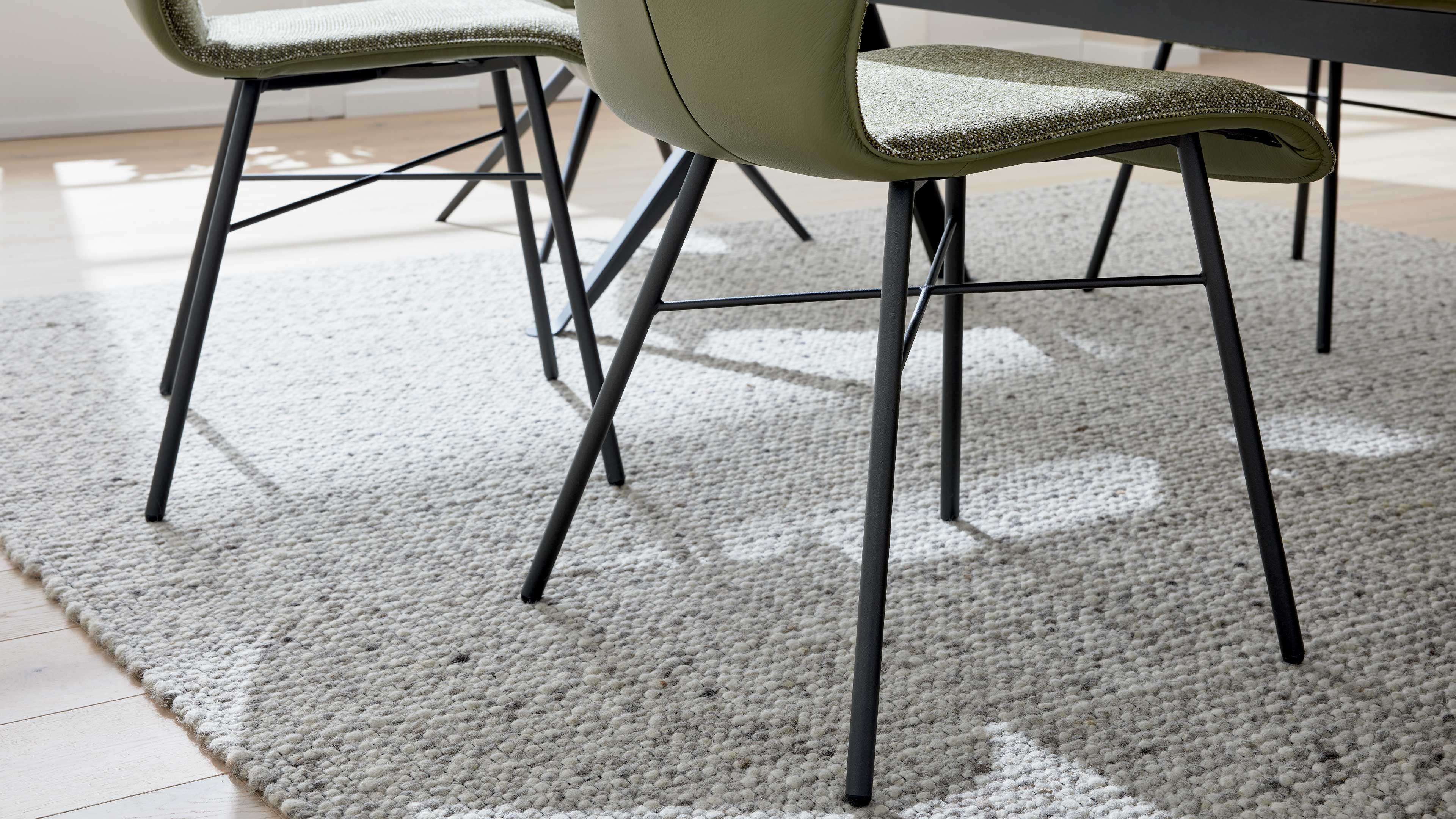 Zwei grün bezogene Stühle mit schwarzen Metallbeinen stehen auf einem hellgrauen, grob gewebten Teppich.