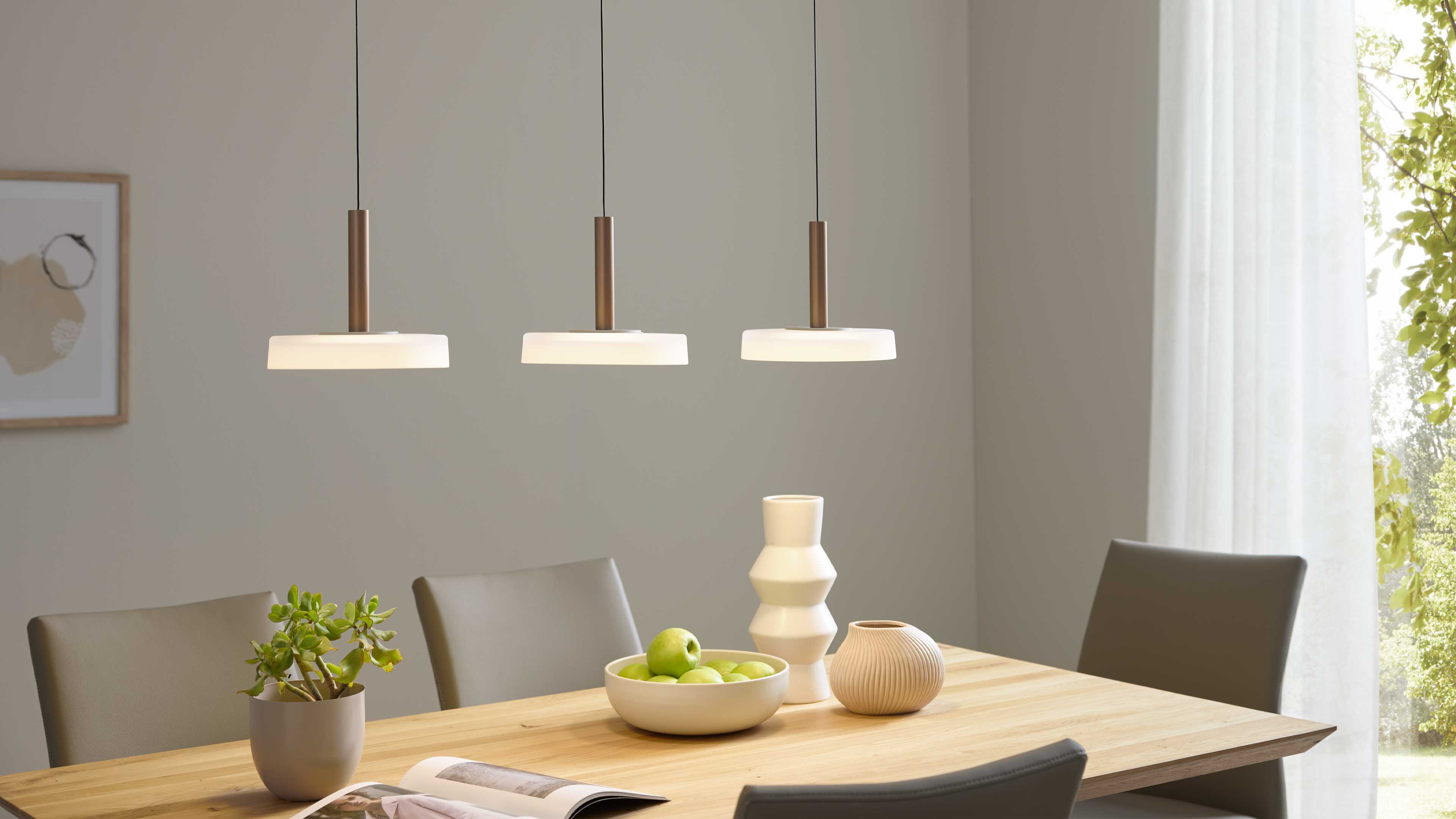 Drei hängende Lampen über einem hellen Holz-Esstisch mit grauen Stühlen, einer Schale grüner Äpfel, Vasen und Topfpflanze.