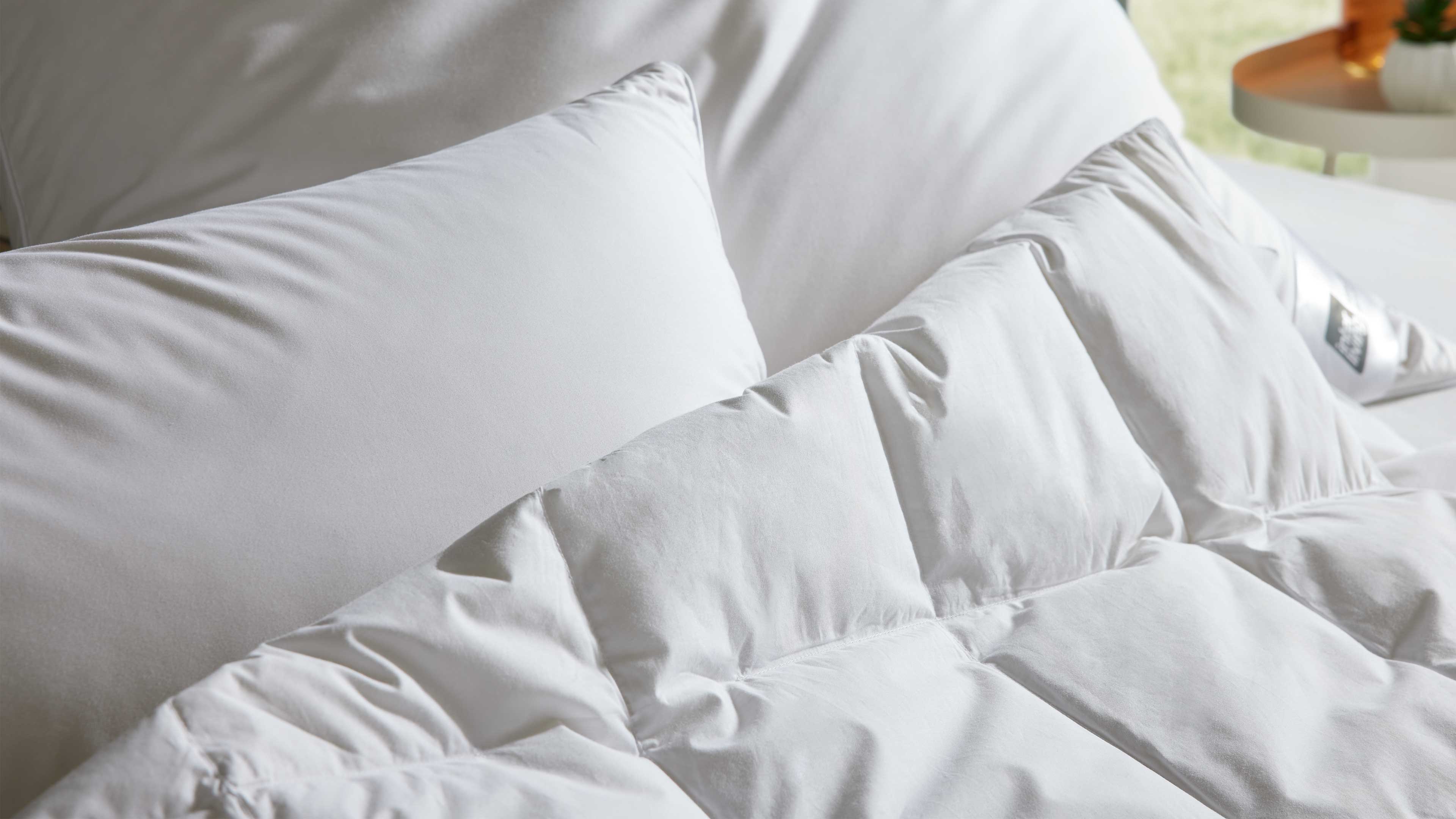 Zwei weiße Kopfkissen und eine gesteppt wirkende weiße Steppdecke auf einem Bett, rechts ein Beistelltisch.