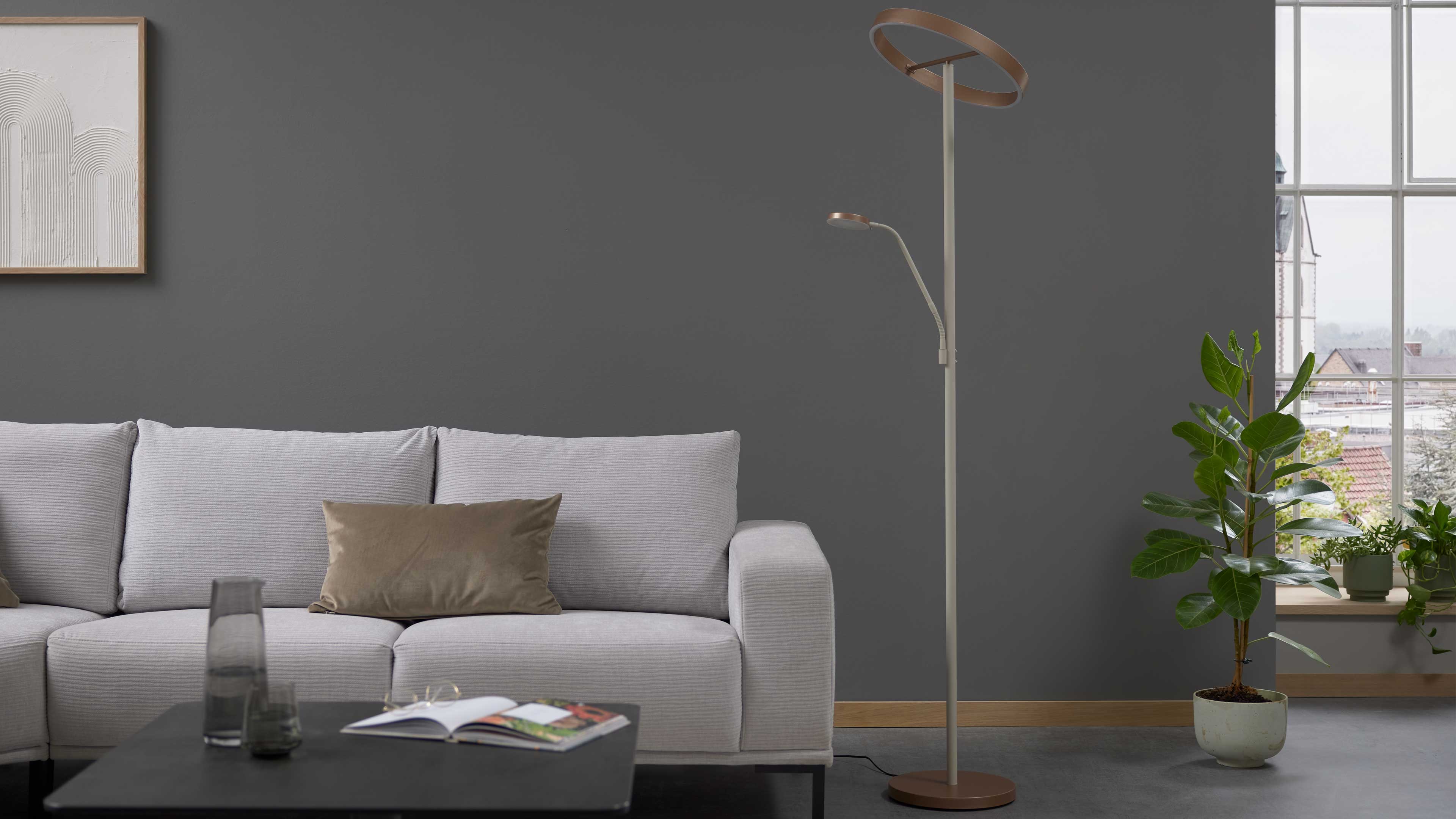 Hellgraues Stoffsofa mit braunem Kissen vor dunkler Wand, daneben schlanke Stehleuchte mit Ringkopf und Pflanze am Fenster.