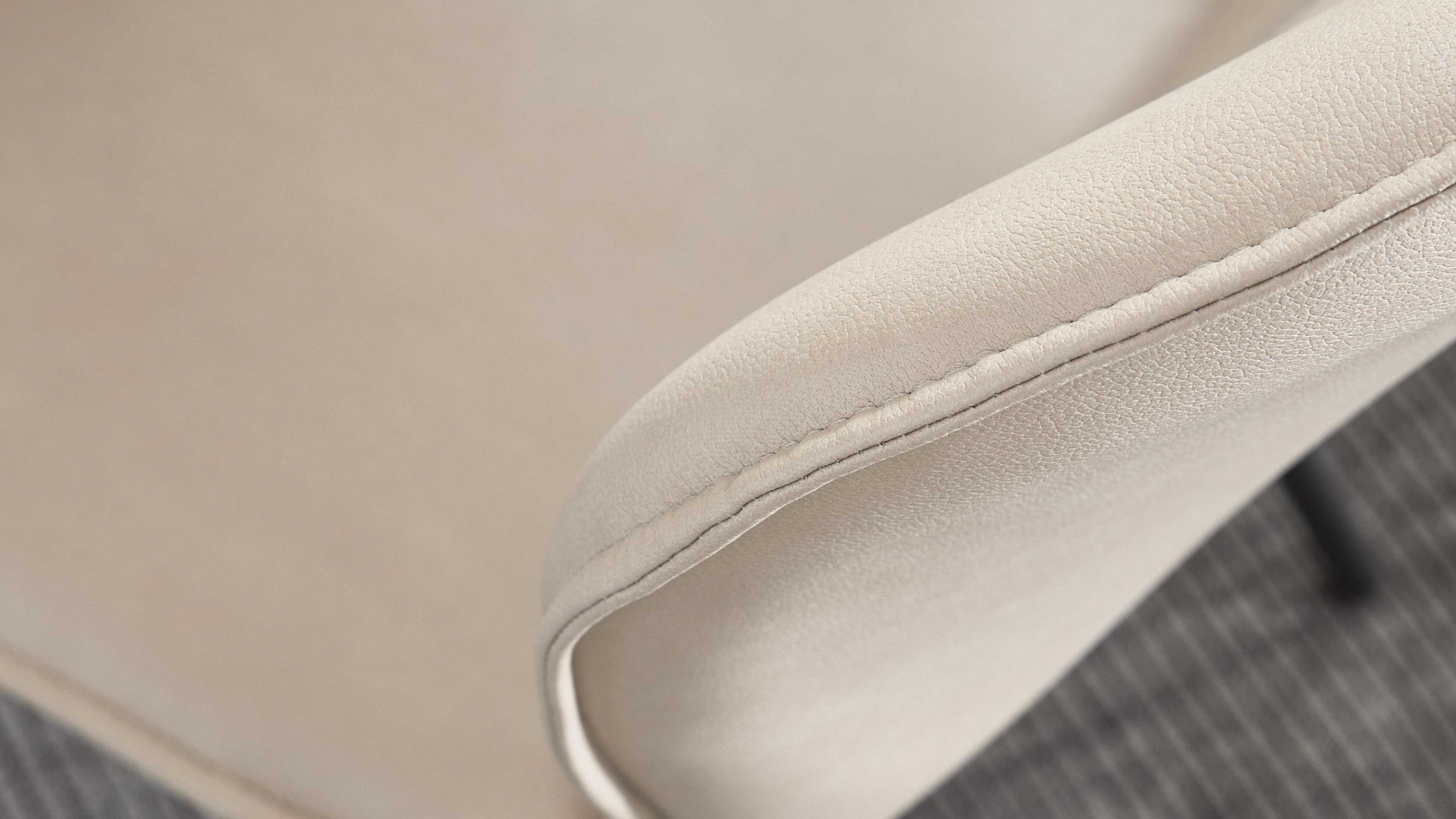 Nahaufnahme einer hellbeigen, leicht genarbten Armlehne mit sichtbarer Naht, vor unscharfem Sitzpolster und Teppichboden.