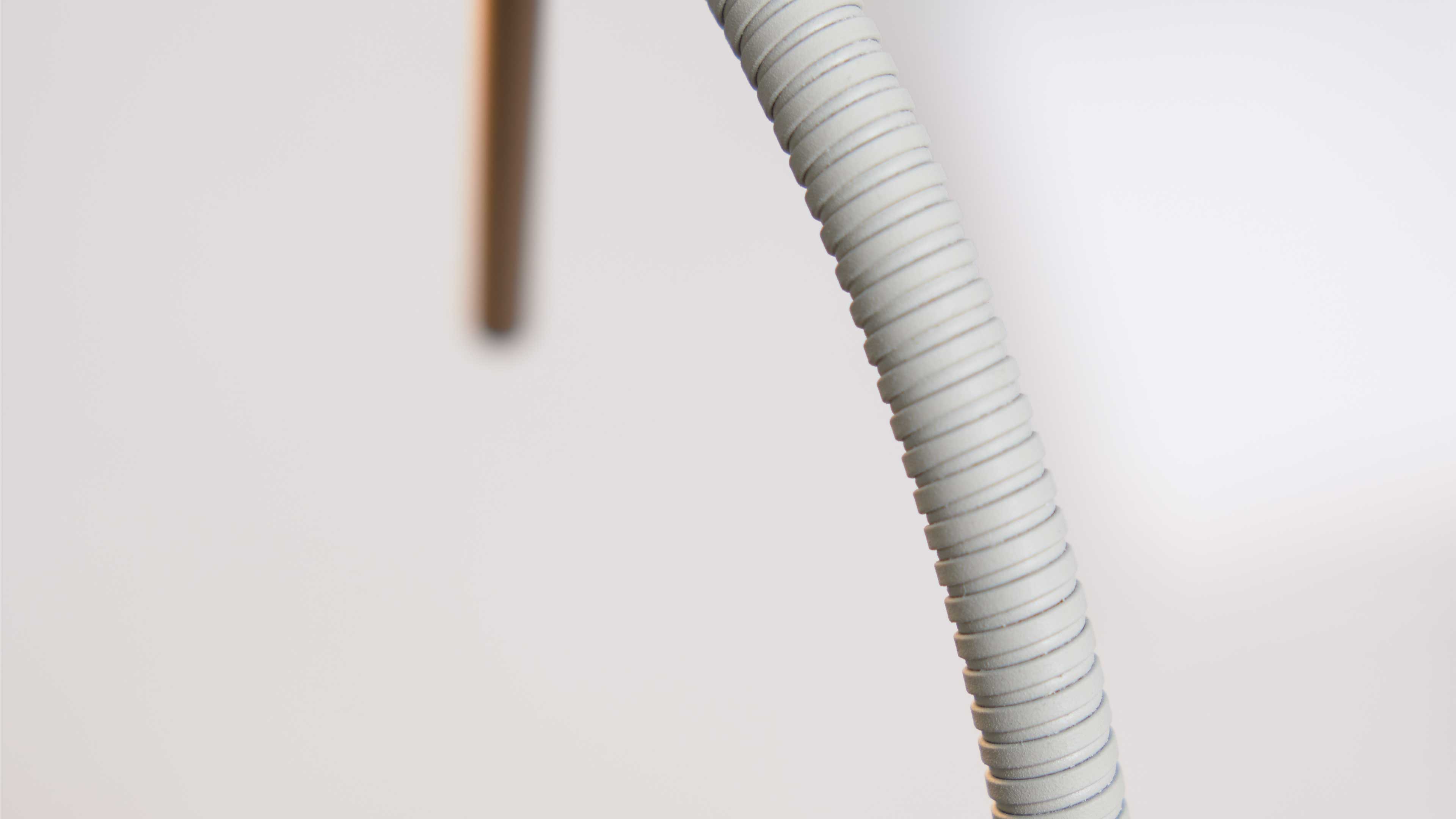 Gebogener, gerippter hellgrauer Kunststoffschlauch mit spiralförmigen Rillen im Vordergrund, unscharfer Holzstab im Hintergrund.