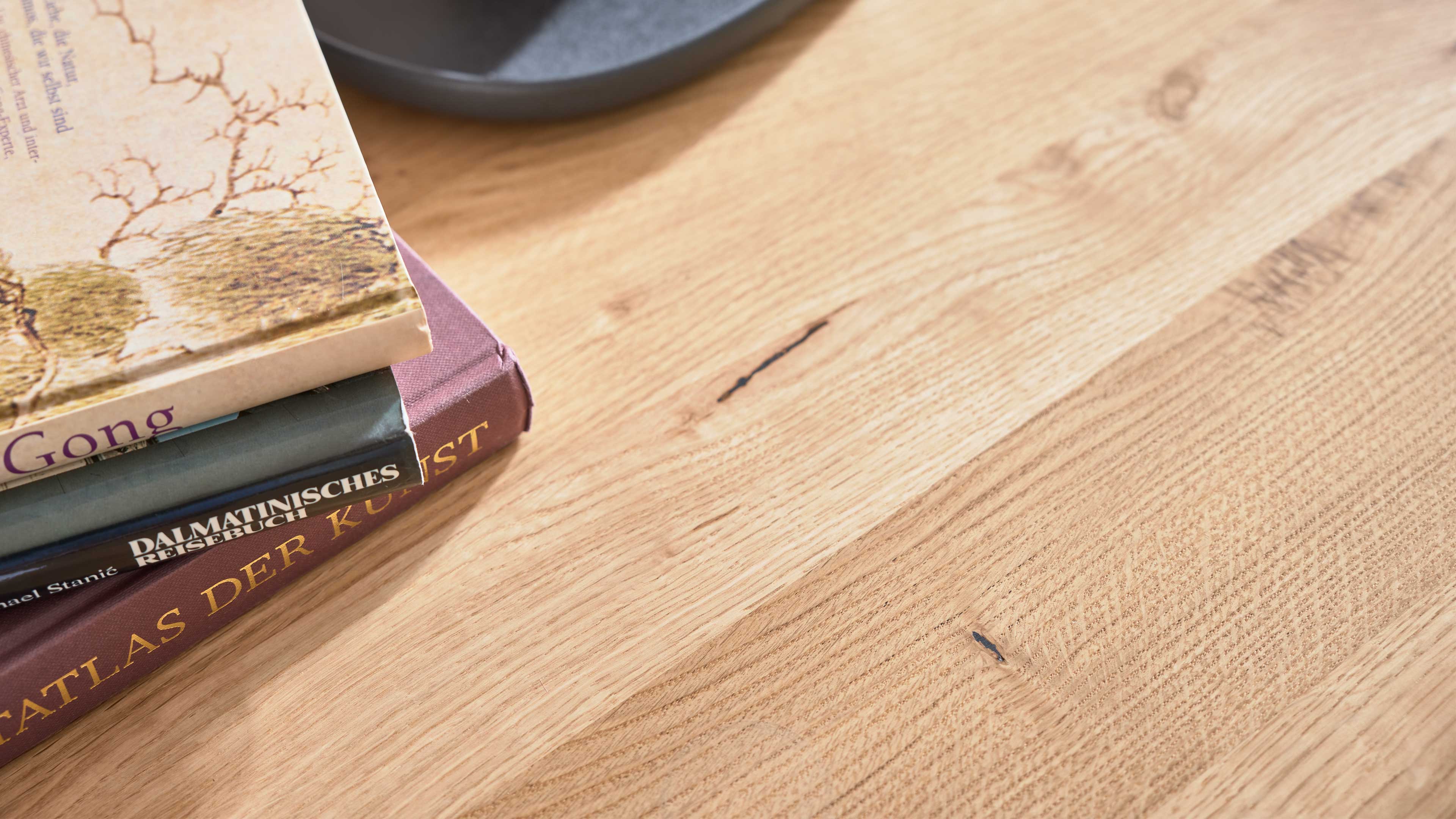 Heller Holztisch mit ausgeprägter Maserung und dunklen Ast- und Rissstellen, links ein Stapel Bücher und ein dunkler Teller.