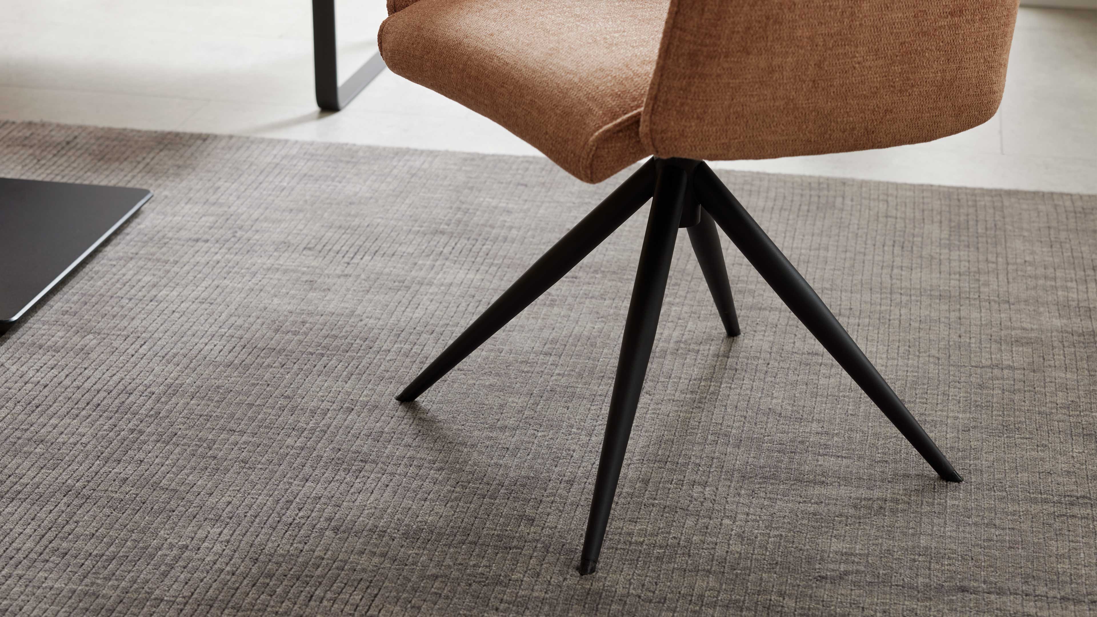 Stuhl mit braunem Stoffsitz und schwarzem vierstrahligem Metallfuß auf grauem, fein geripptem Teppich.