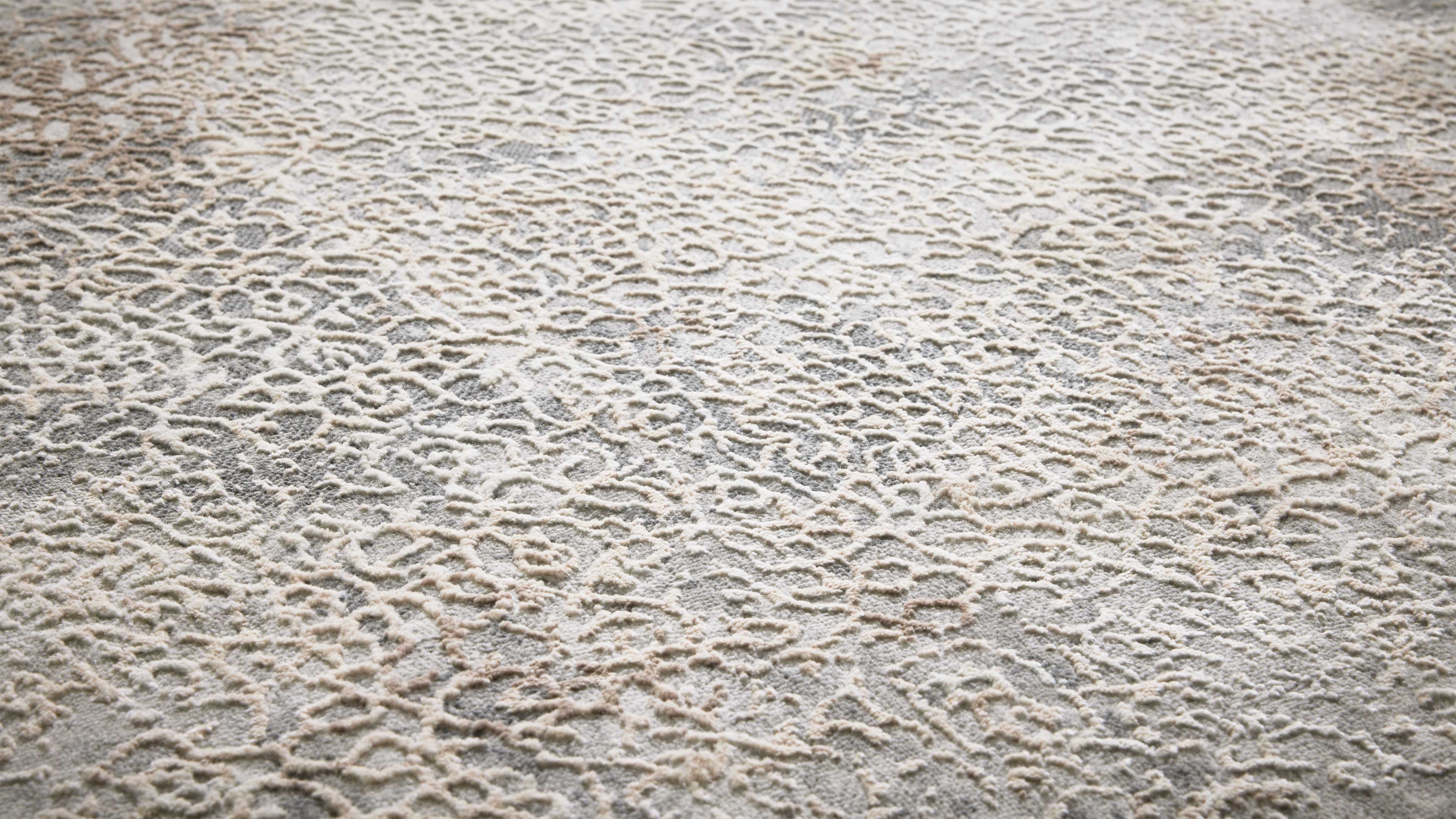 Hellgrauer Teppich mit reliefartigem cremefarbenem Muster aus erhöhten, unregelmäßigen Linien und Schleifen.