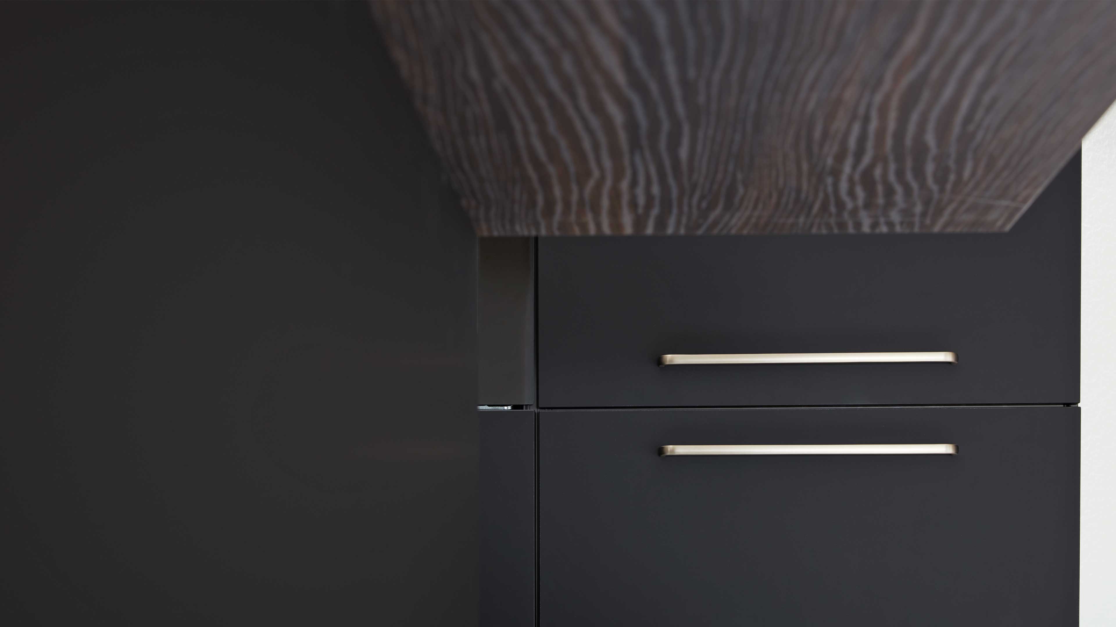 Zwei dunkle matte Küchenschubladen mit schlanken silbernen Griffen, darüber eine dunkle Holzplatte mit deutlicher Maserung.