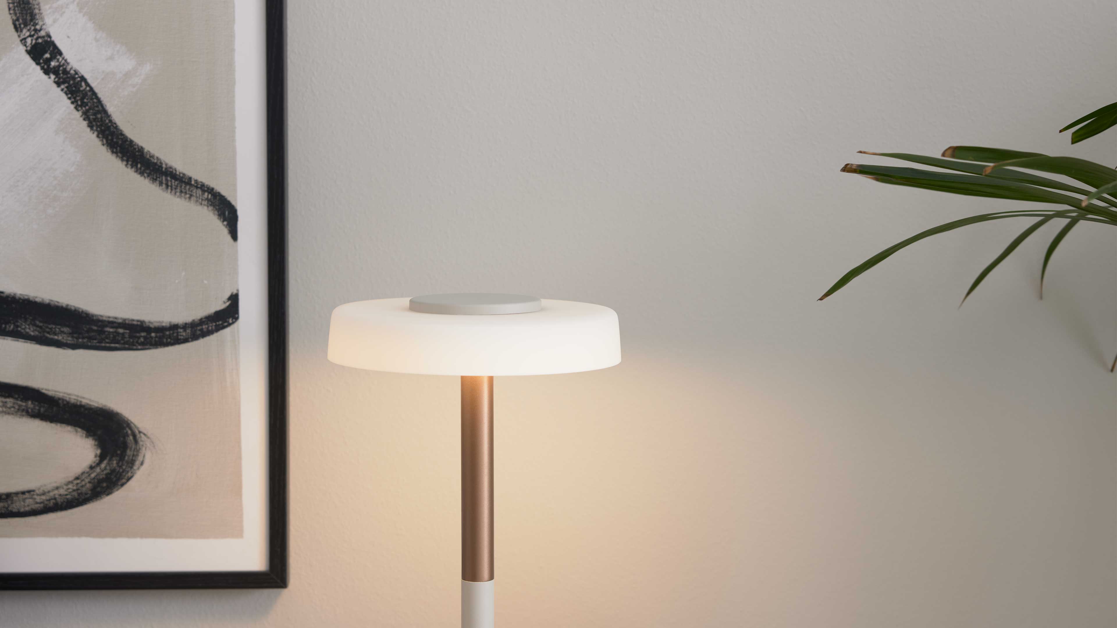 Runder weißer Lampenschirm auf dünnem kupferfarbenem Stab, links gerahmtes schwarz-weißes Pinselbild, rechts grüne Blätter.