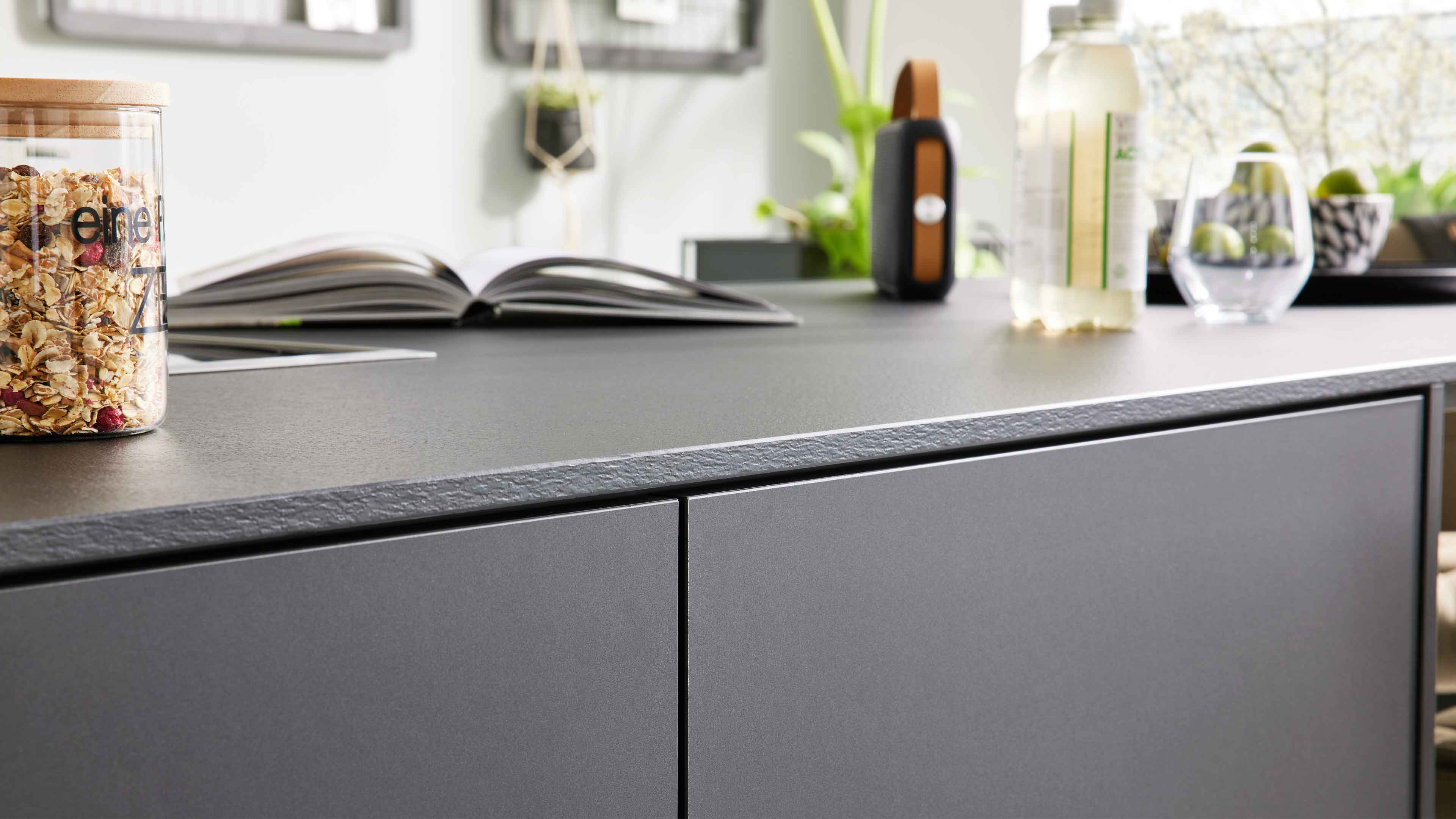 Dunkelgraue Küchenfront mit strukturierter Arbeitsplatte, darauf ein Glasbehälter mit Müsli, ein geöffnetes Buch, Flasche und Glas.