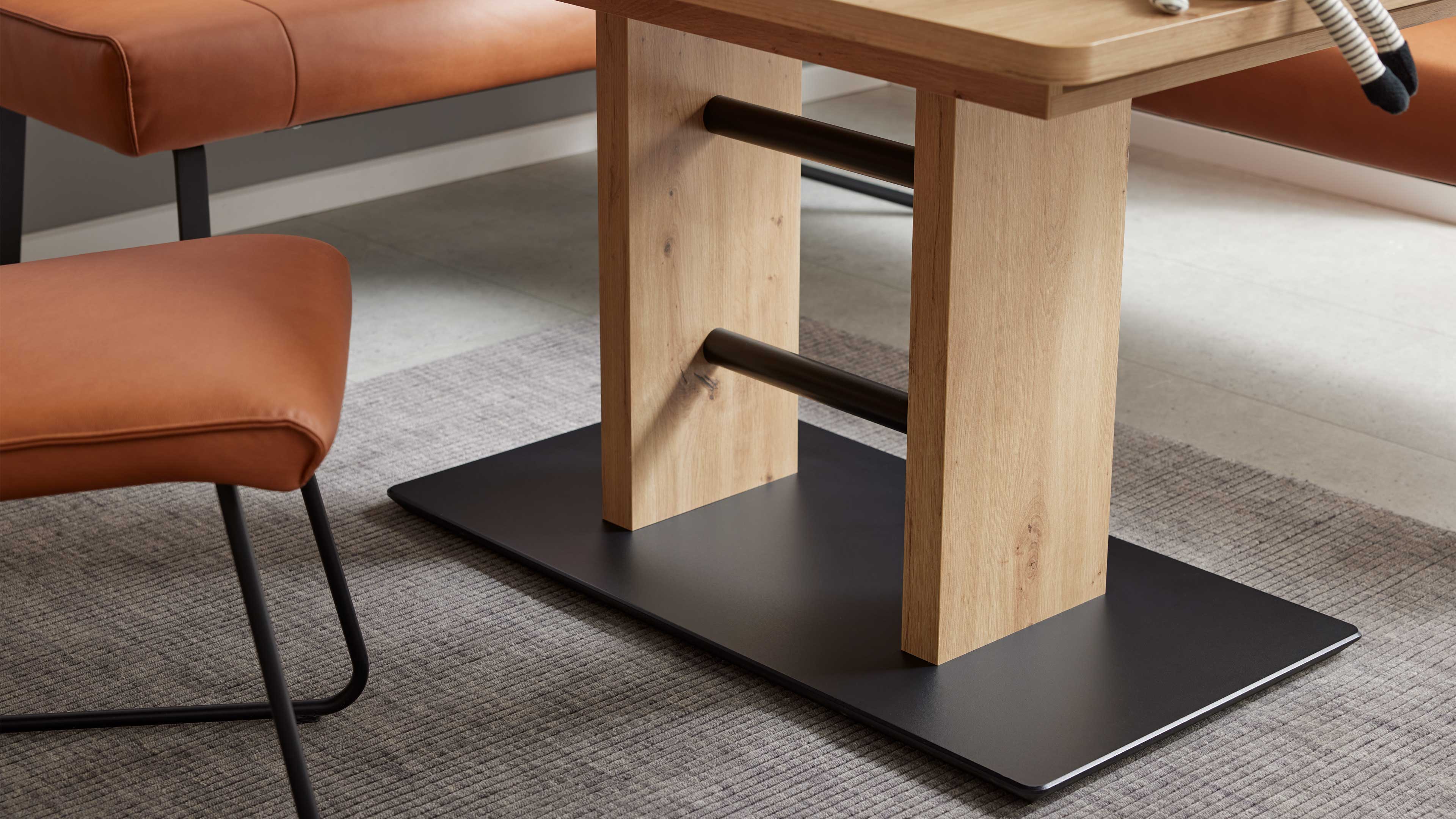 Tischgestell mit zwei hellen Holzsäulen, schwarzen Metallstreben und schwarzer Bodenplatte, daneben braune Polster auf schwarzen Füßen.