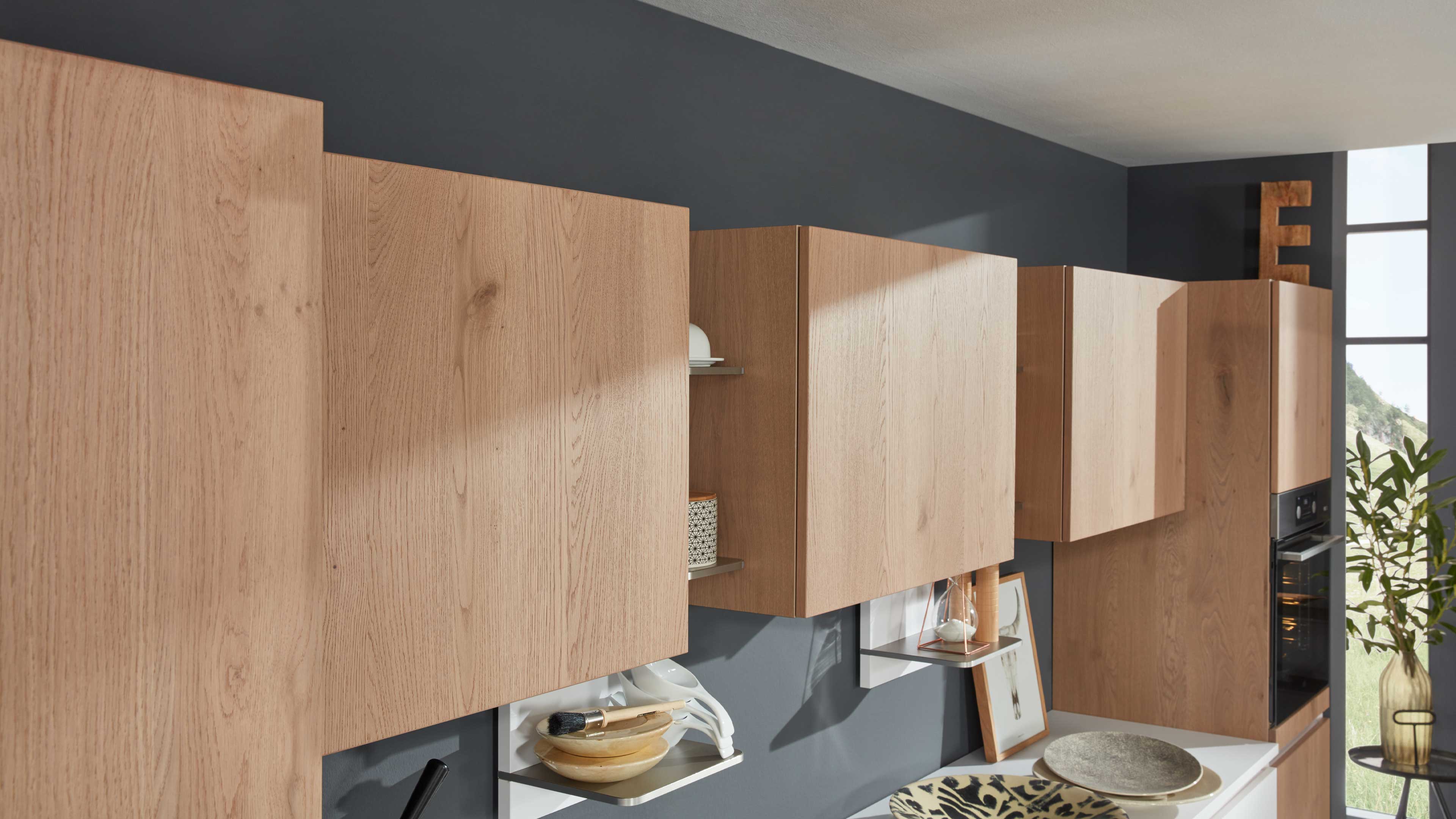 Reihe hängender Küchenoberschränke mit hellen Eichenoptikfronten und vertikaler Maserung vor dunkelgrauer Wand, teils offen mit Ablagen.