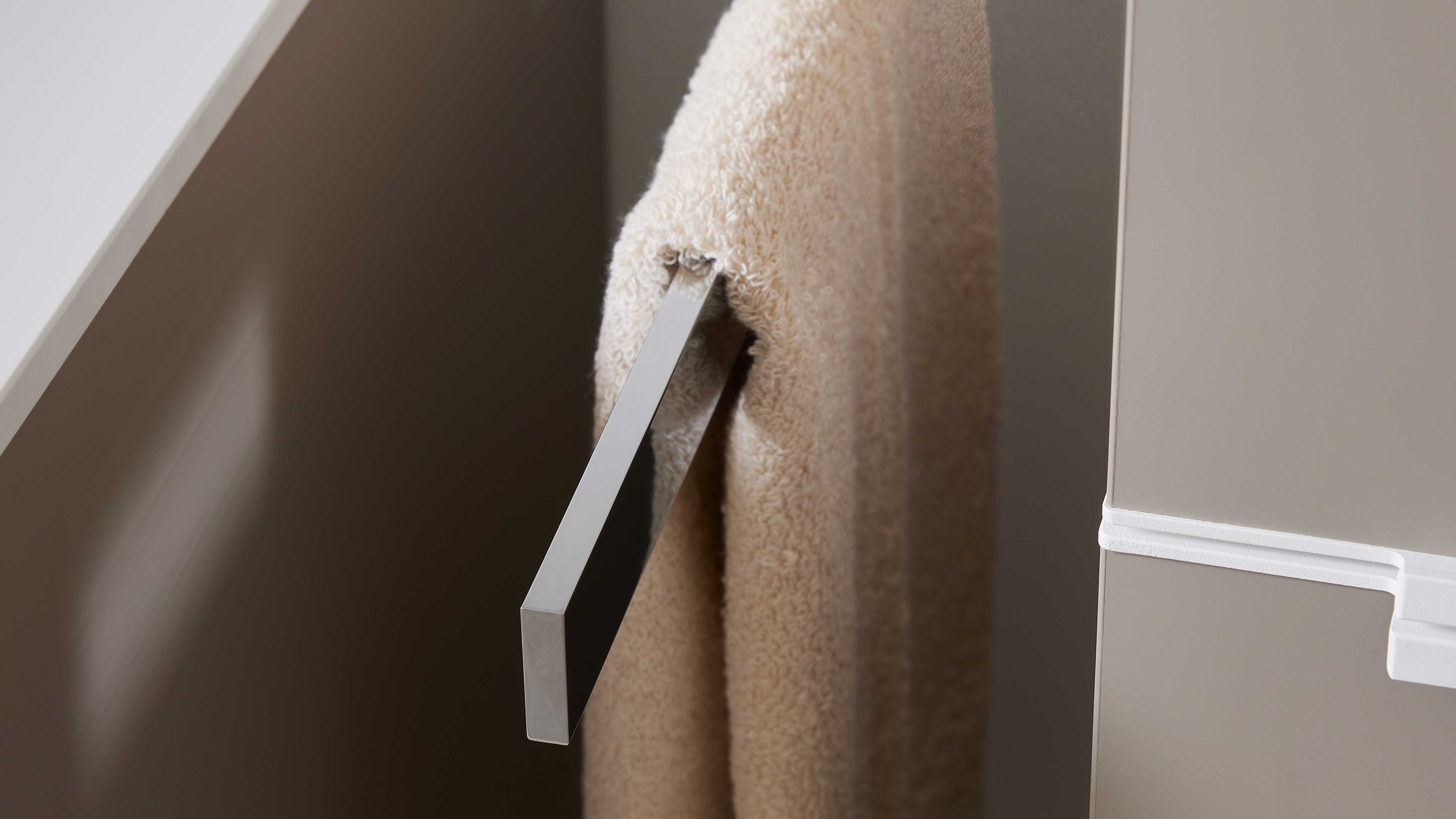 Flache, rechteckige Metallstange mit einem beigen Frotteehandtuch, herausgezogen aus einer hellgrauen Schrankfront.