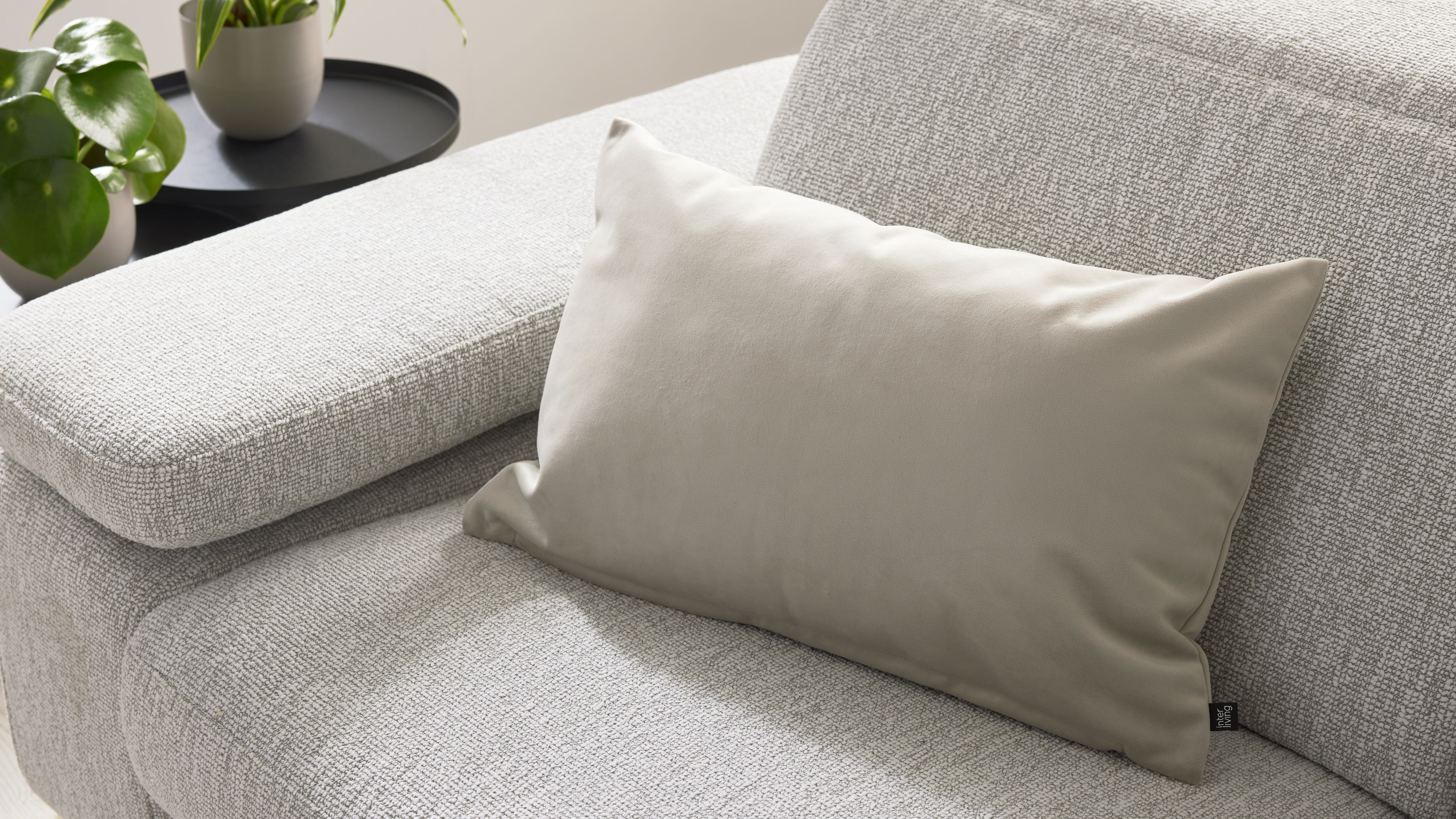 Beiges rechteckiges Kissen liegt auf hellgrauem, strukturiertem Sofa; links ein schwarzer Beistelltisch mit zwei Pflanzen.