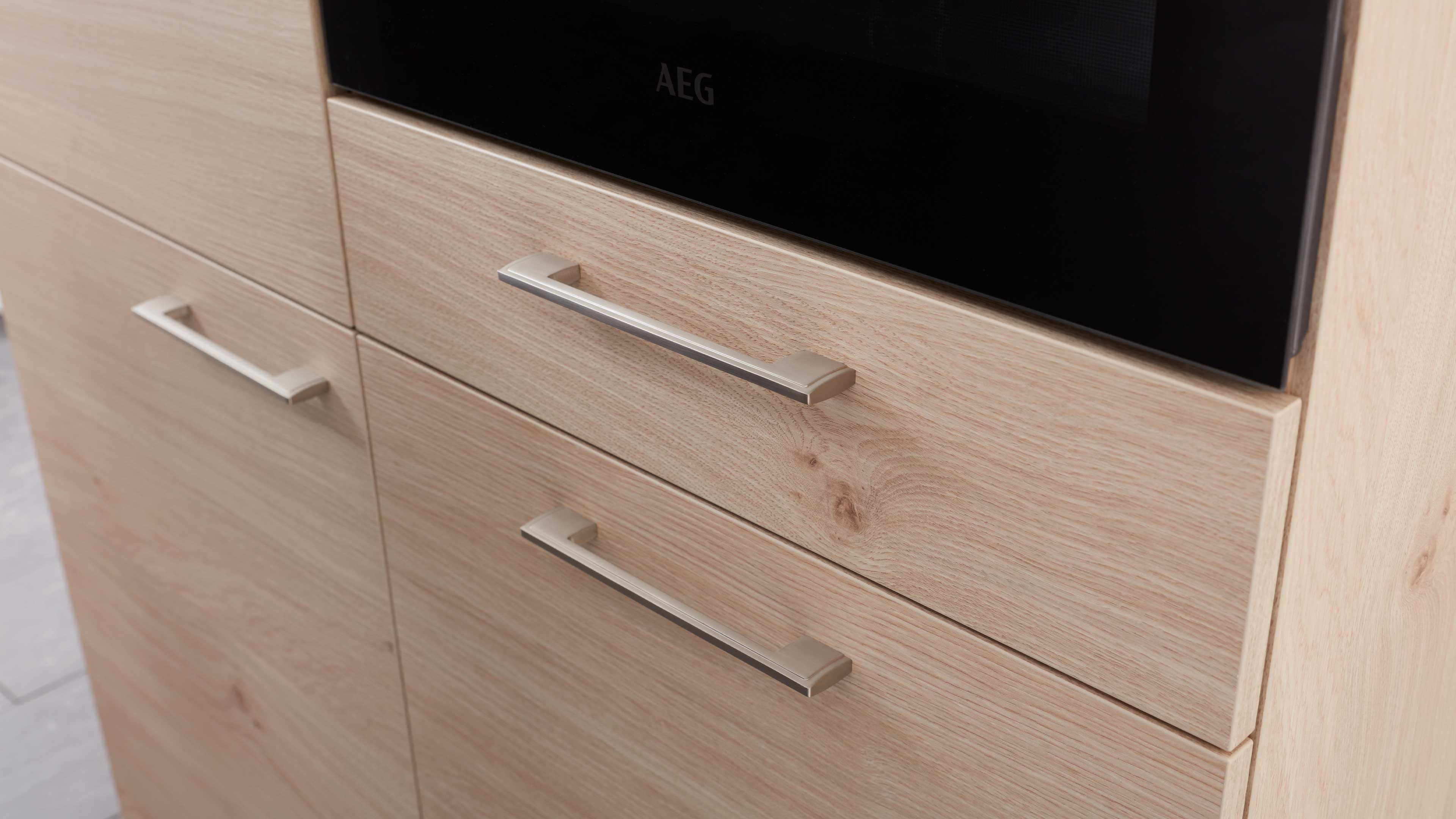 Helle eichenfarbene Schrank- und Schubladenfronten mit sichtbarer Holzmaserung und langen, flachen Metallgriffen unter schwarzer Ofenfront.
