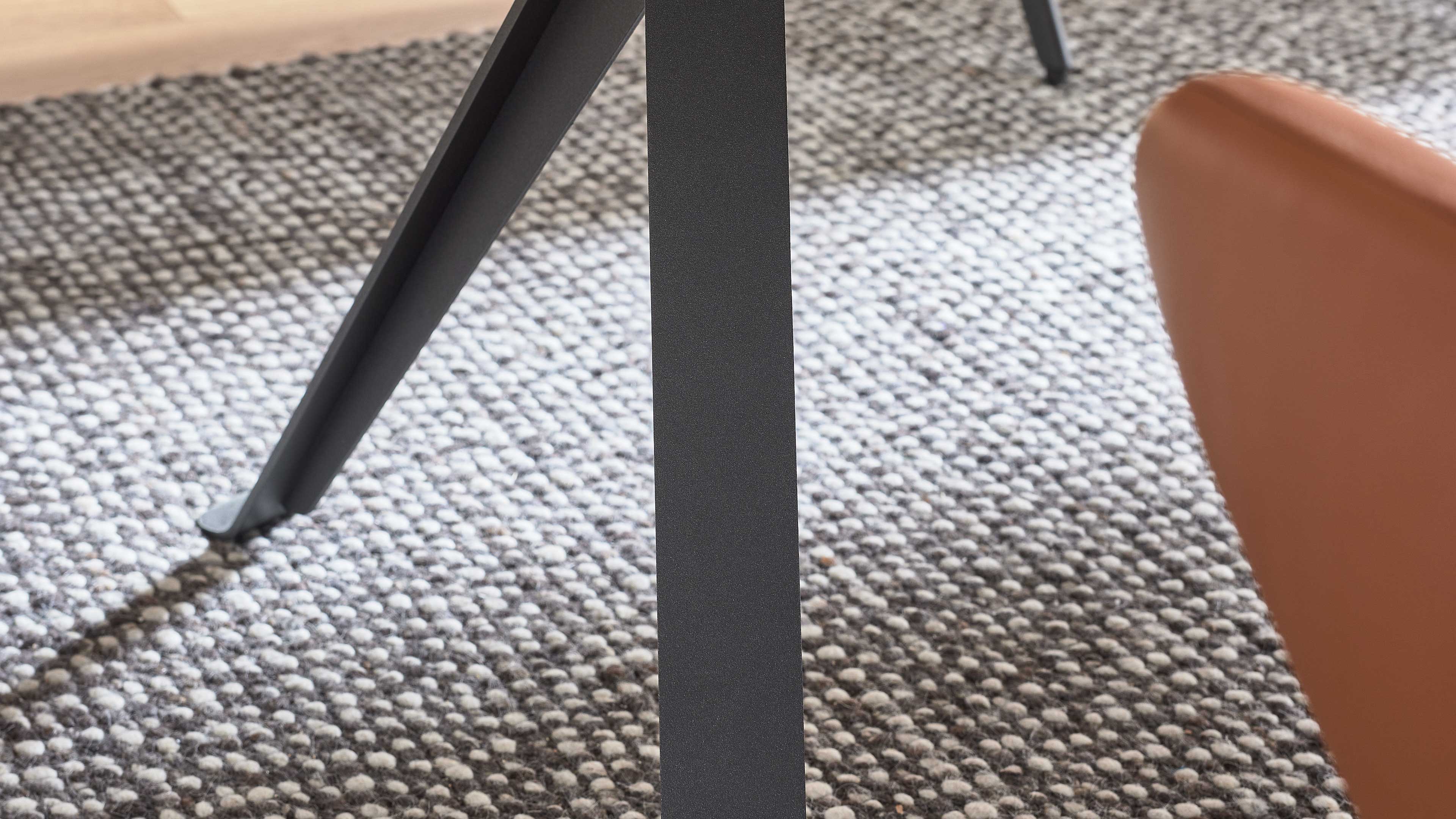 Mittiges schwarzes Metall-Tischbein und links diagonales Stützbein, rechts braune Sitzlehne auf grob gewebtem grauem Teppich.