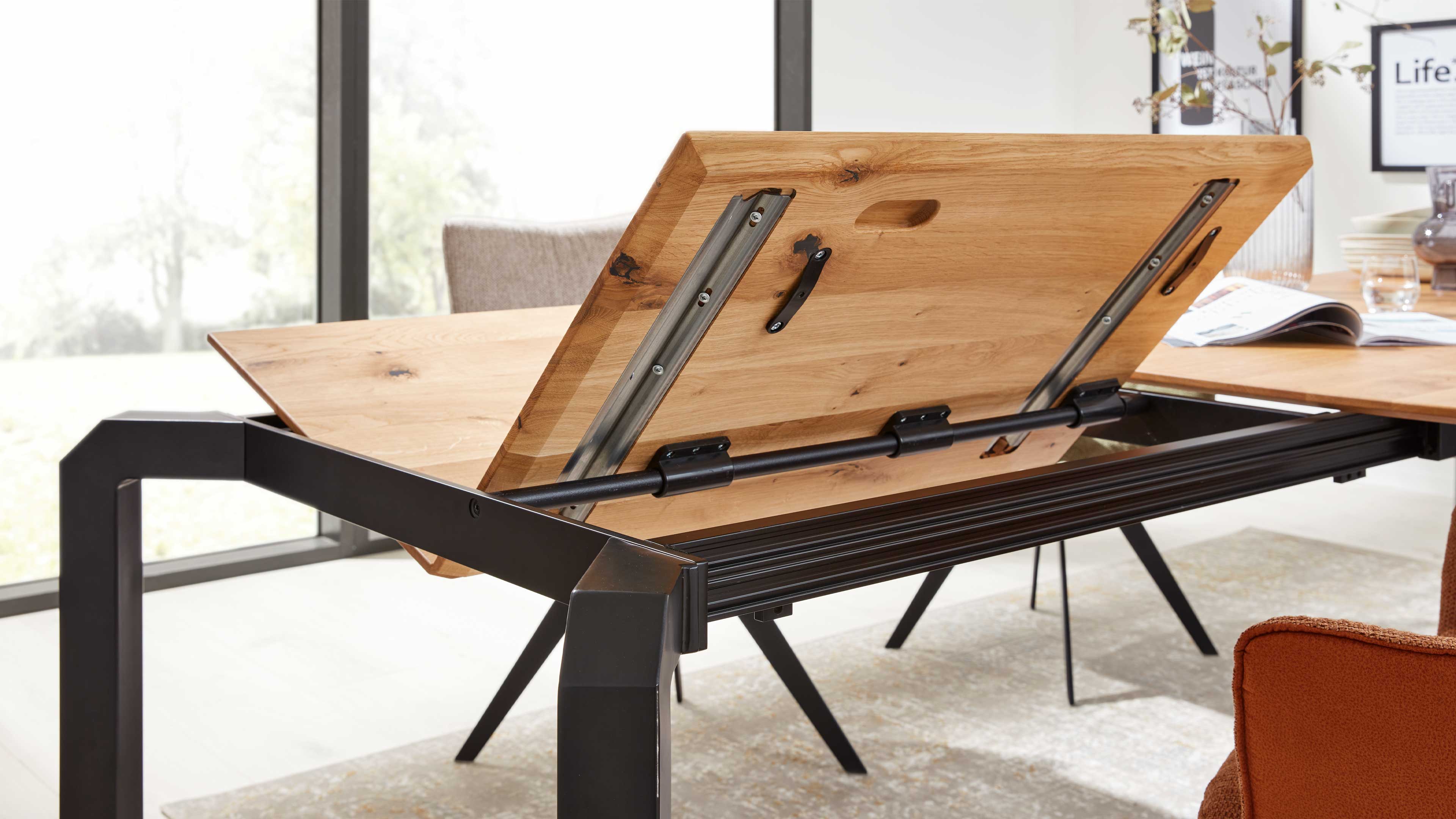 Ausgeklappte Holz-Tischplatte mit sichtbaren Metallauszügen und schwarzem Gestell, im Hintergrund Fenster und orangefarbener Sessel.