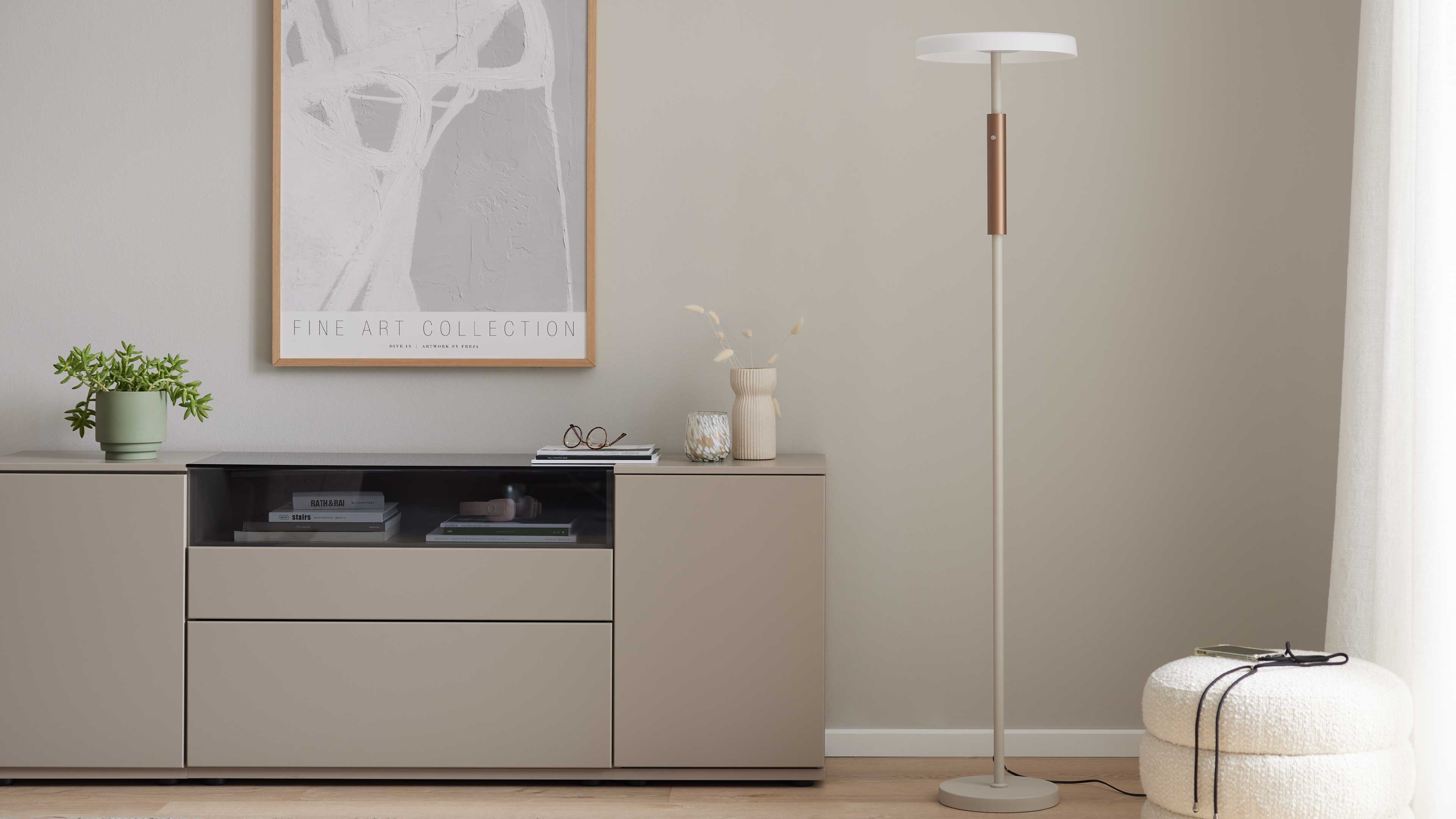 Beiges Sideboard mit offenem Glasfach, Pflanzen, Büchern, Vase und Brille, rechts eine schlanke Stehlampe und ein runder weißer Pouf.