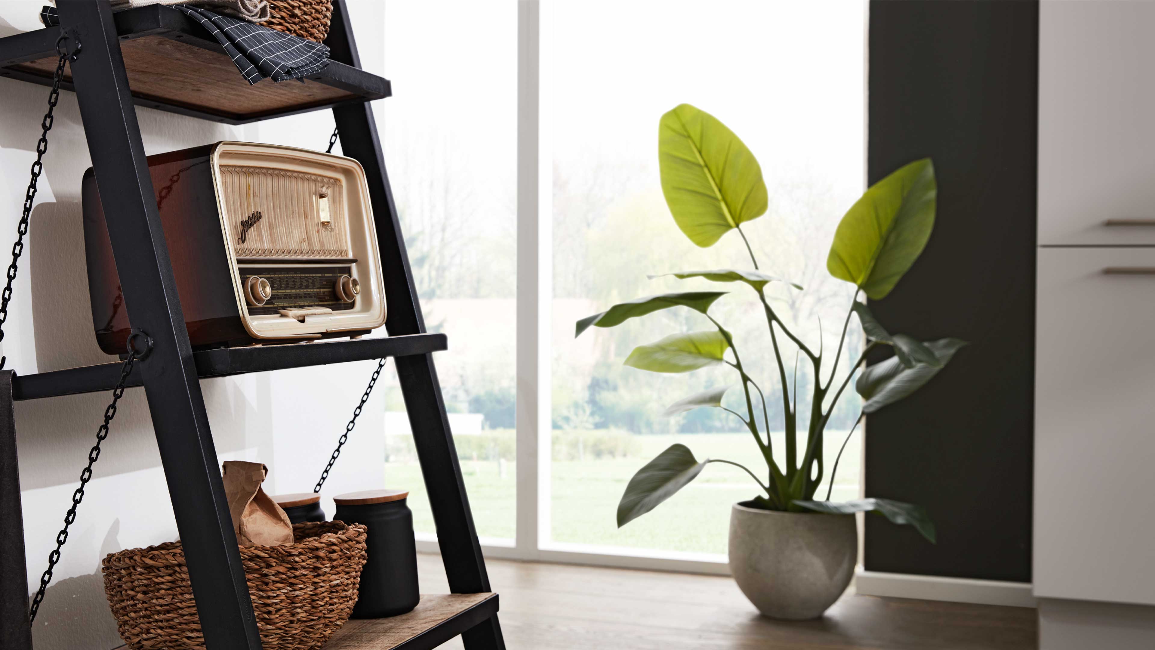 Schwarzes Leiterregal mit altem Radio auf Ablage, darunter Rattankorb und schwarze Vorratsdosen; rechts große Topfpflanze vor Fenster.