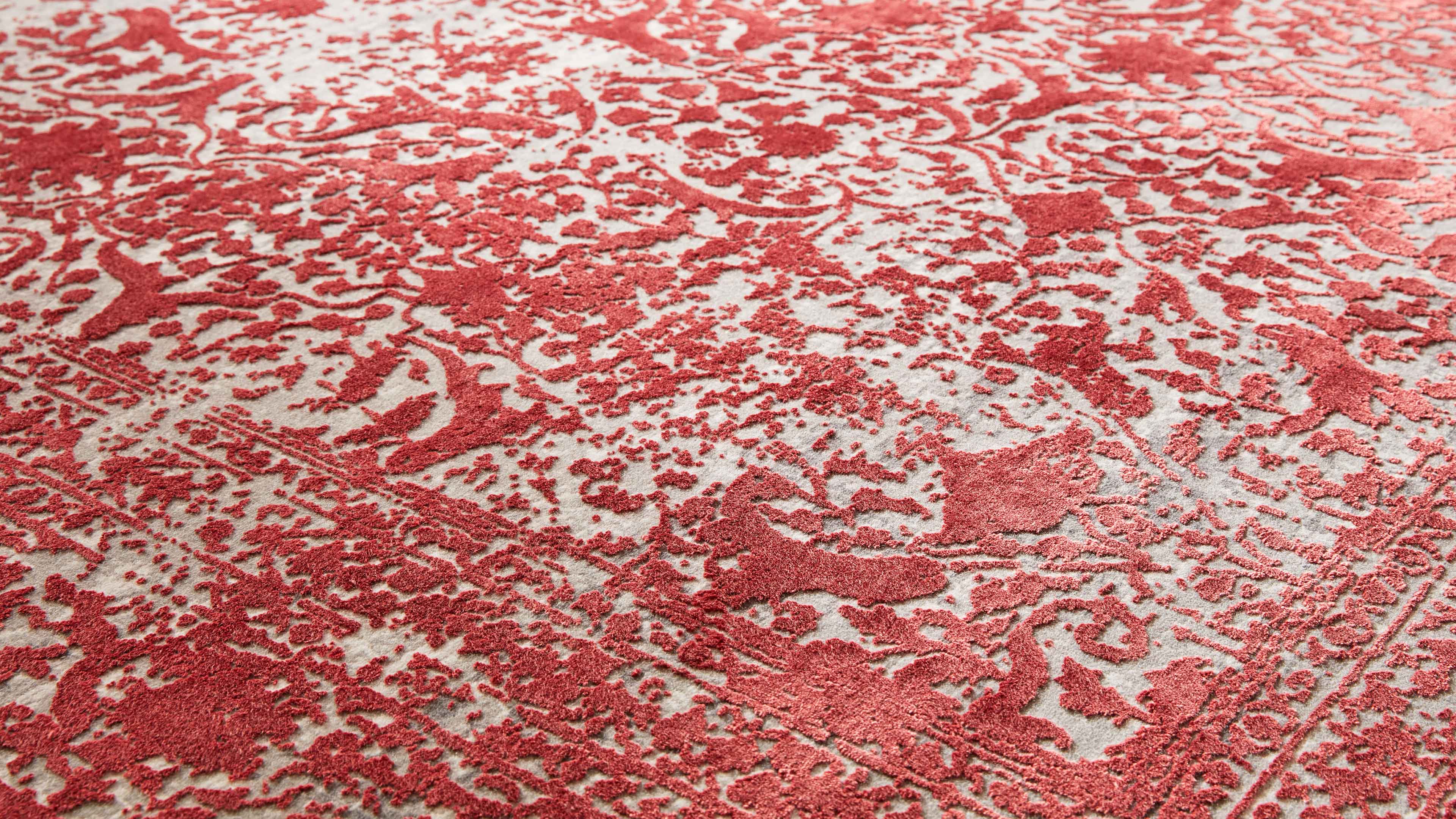 Großaufnahme eines Teppichs mit hellbeigem Grund und erhabener, roter Ornament- und Blumenmusterung samt Randlinie.