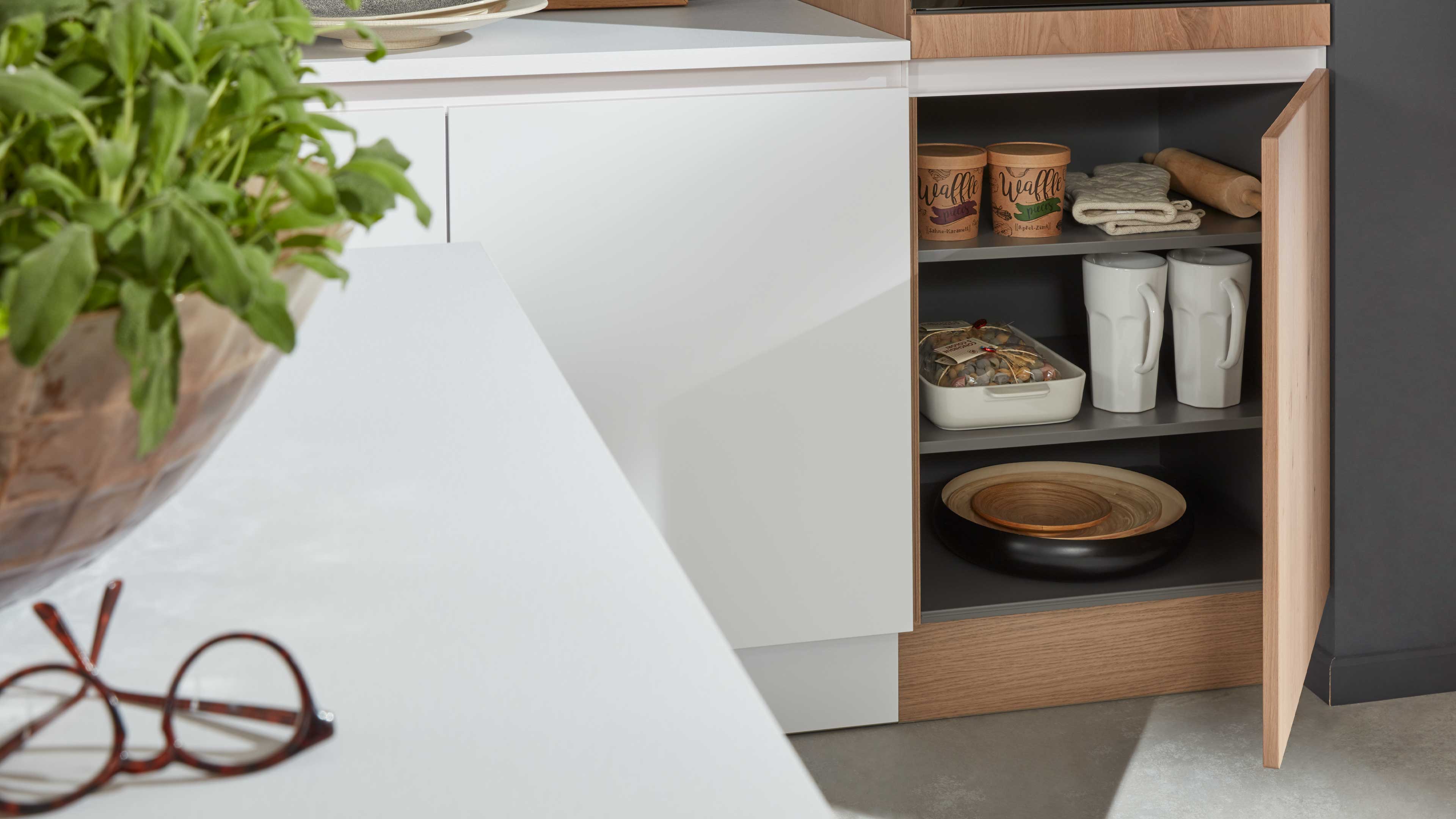 Offener Küchenunterschrank mit weißer Front und Holzrahmen, grauen Einlegeböden mit Tassen, Schalen, Dosen und Textilien.