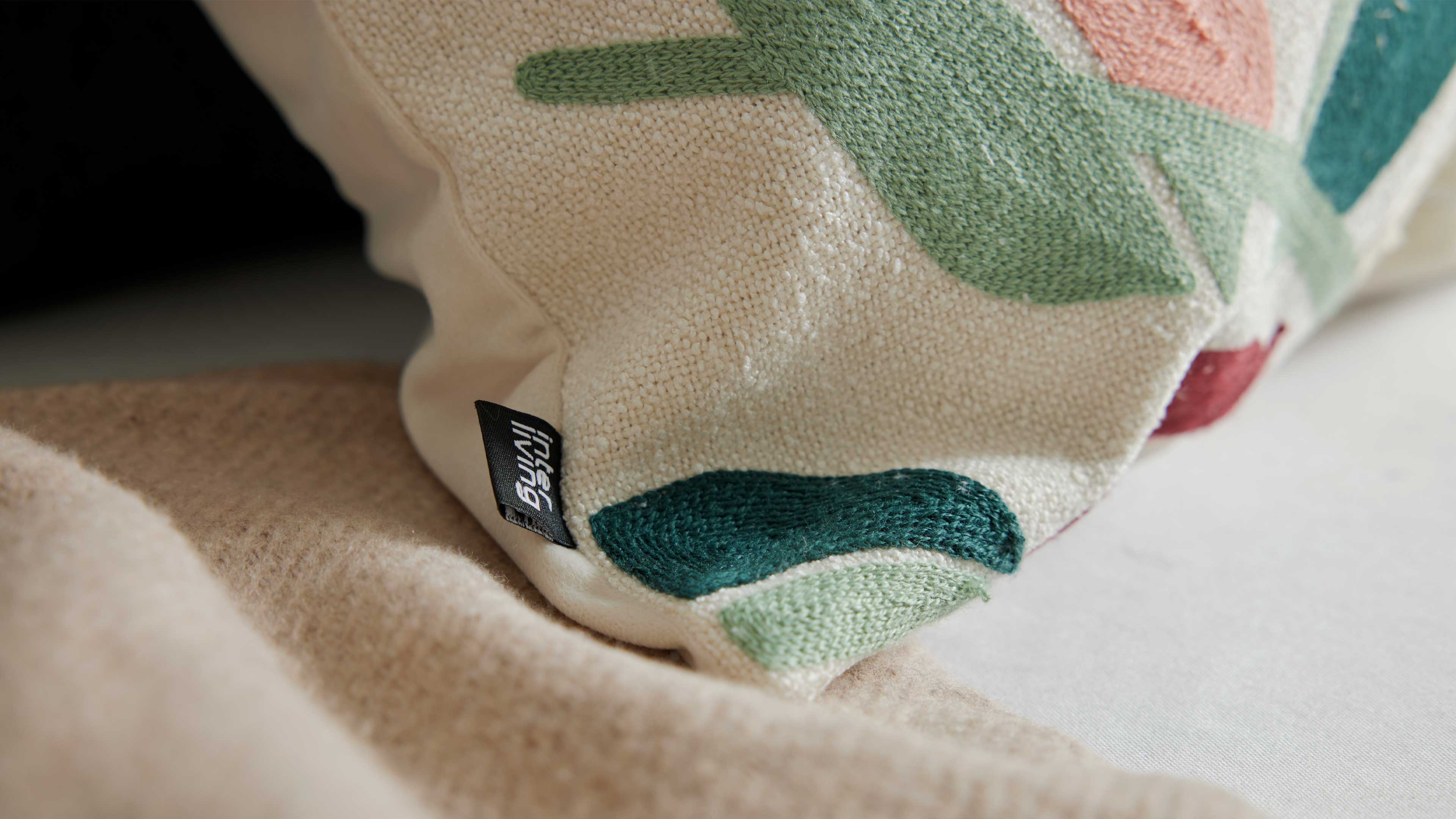 Nahaufnahme eines ecrufarbenen Kissenecks mit gestickten grünen und rosa Formen, sichtbarem schwarzen Textil-Etikett und beiger Decke.