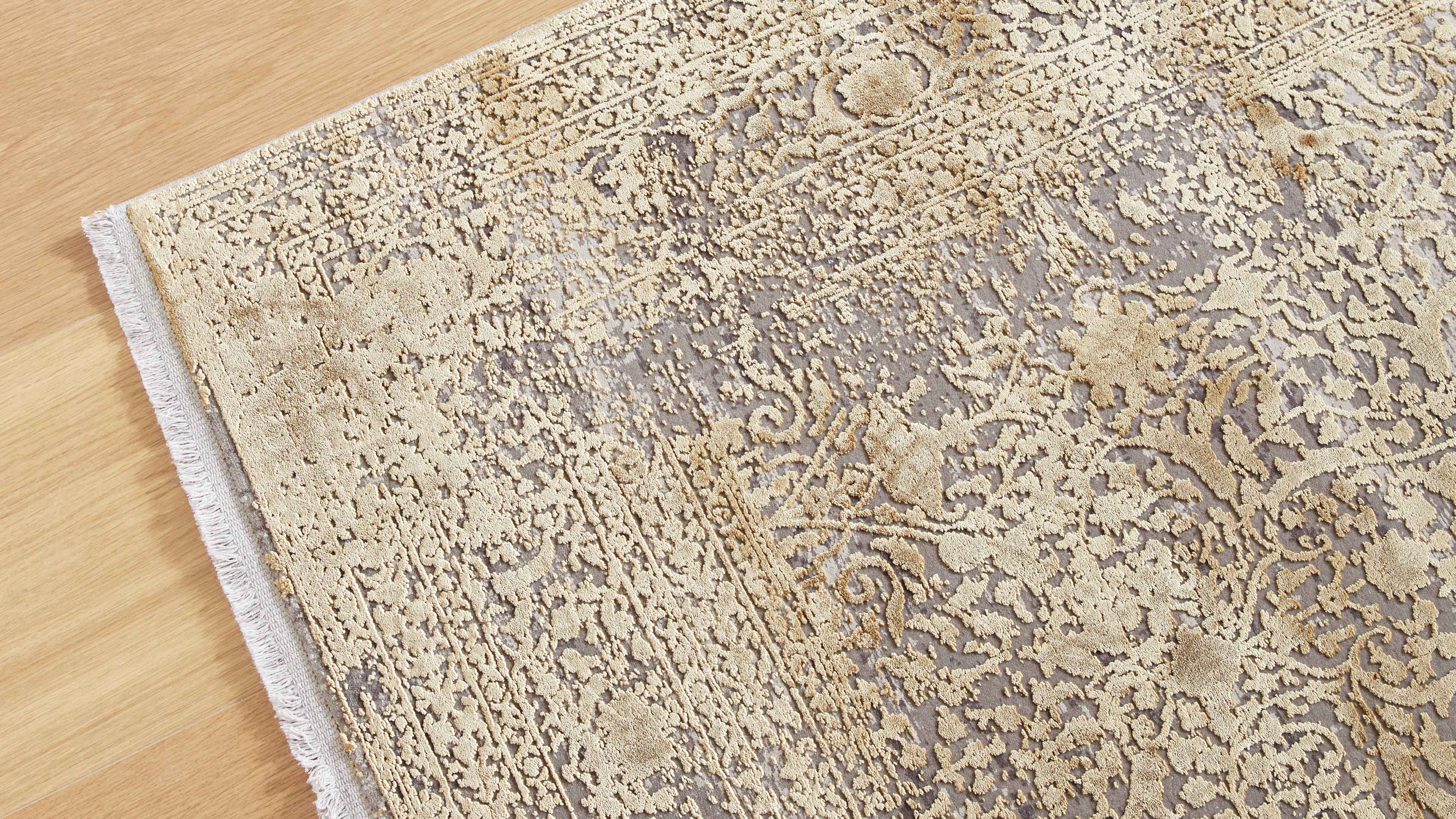 Teppich mit beigem, reliefartigem Ornamentmuster auf graubraunem Grund, sichtbare Fransen links und heller Holzboden.
