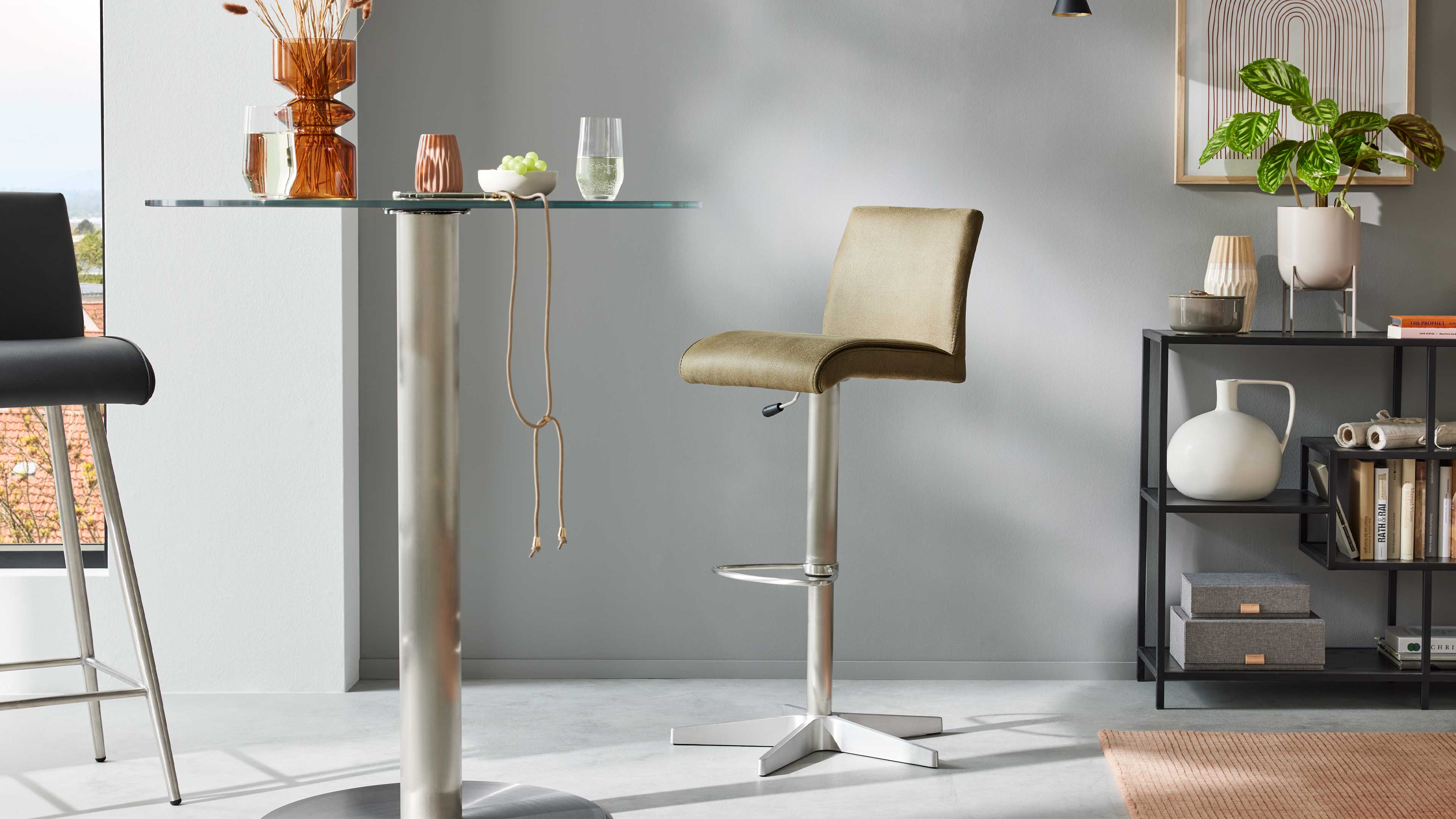 Olivegrüner Barhocker mit niedriger Rückenlehne und verchromtem Fuß vor einer grauen Wand, links Glastisch mit bernsteinfarbener Vase.