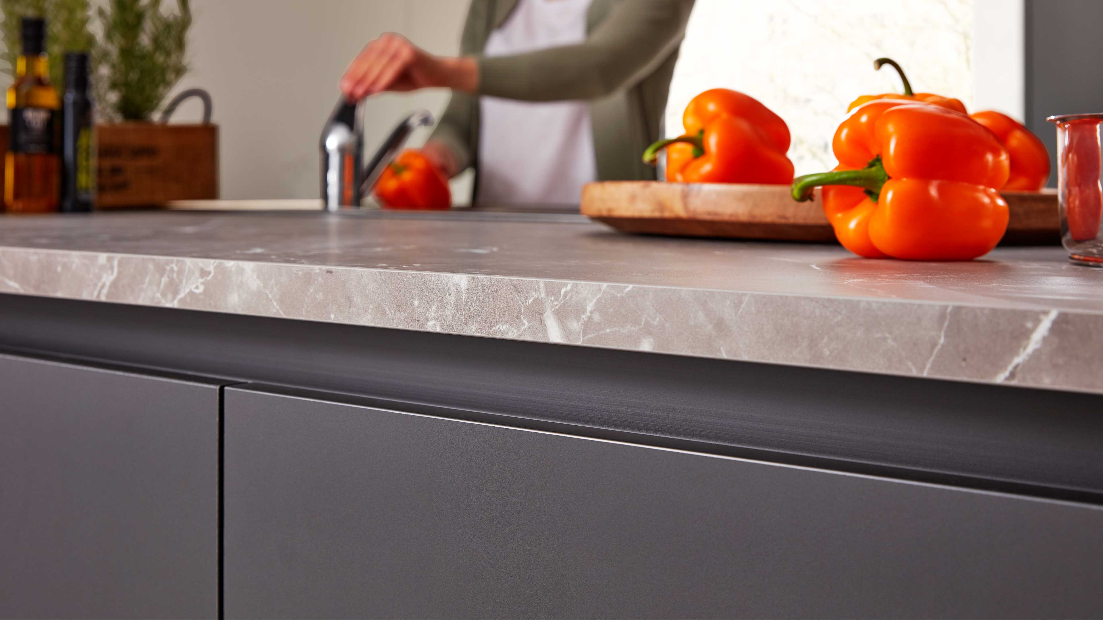 Graue Marmorarbeitsplatte mit Kante über dunklen grifflosen Schrankfronten; im Hintergrund Paprika, Schneidebrett und Wasserhahn.