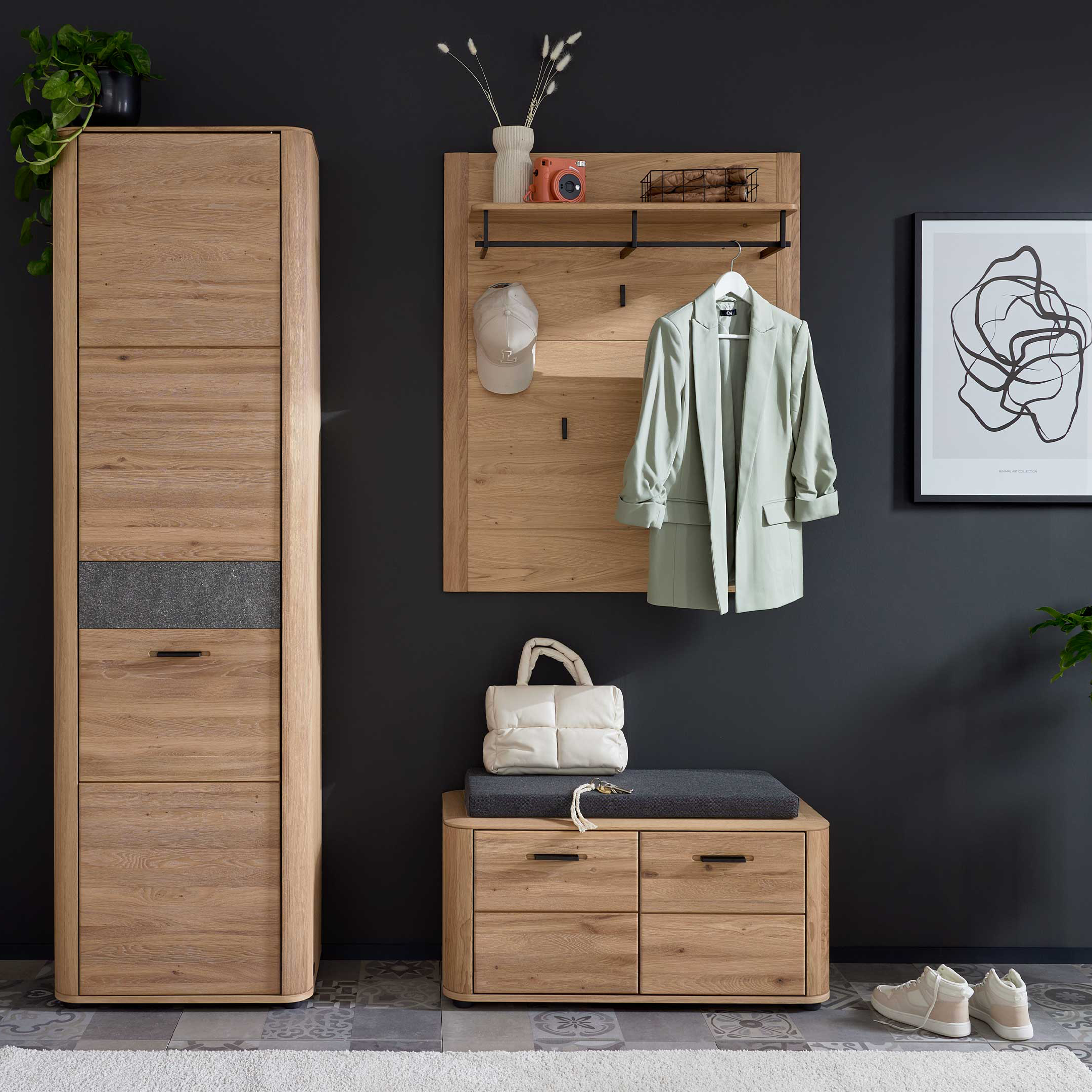 Hoher Holzschrank links, daneben Wandpaneel mit Ablage, hängendem Blazer und Sitzbank, rechts Pflanze und zwei gerahmte Bilder.