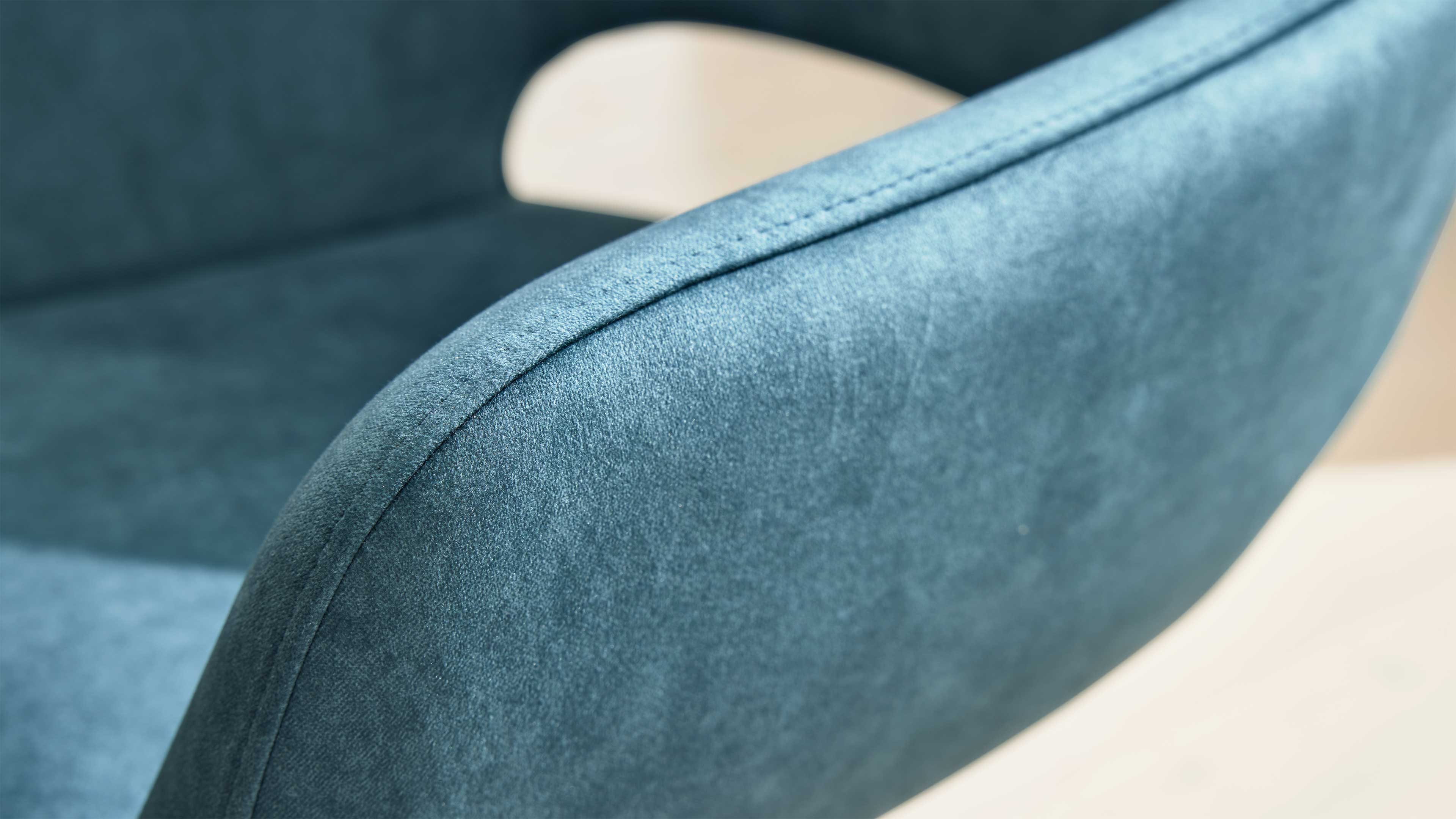 Türkisblaues, samtiges Polster mit sichtbarer Naht an der abgerundeten Armlehne eines Sitzmöbels, heller Boden im Hintergrund.