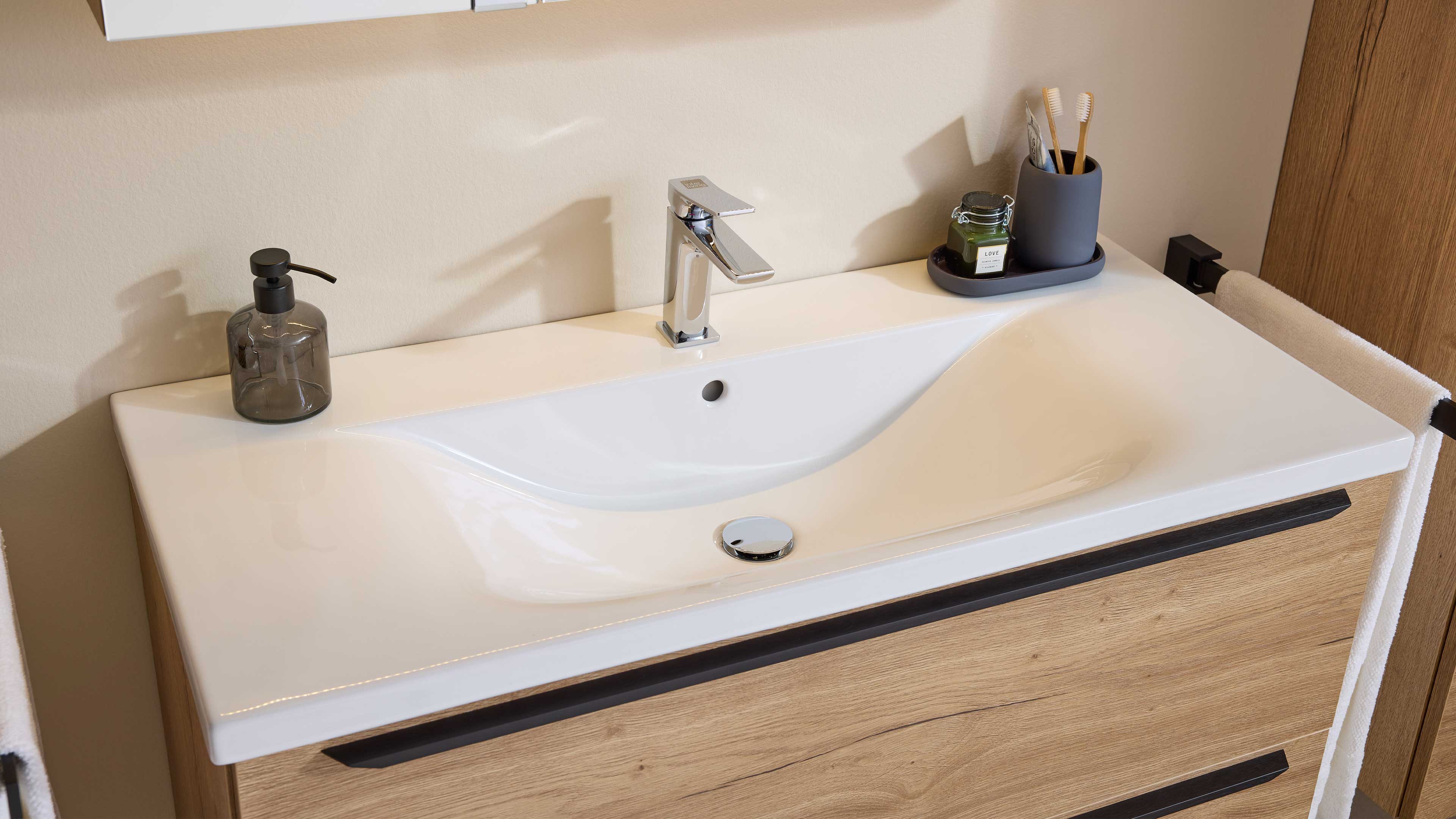 Weißer Keramikwaschtisch mit mittiger Chromarmatur auf Holz-Unterschrank, links Seifenspender, rechts Zahnputzbecher und Handtuch.
