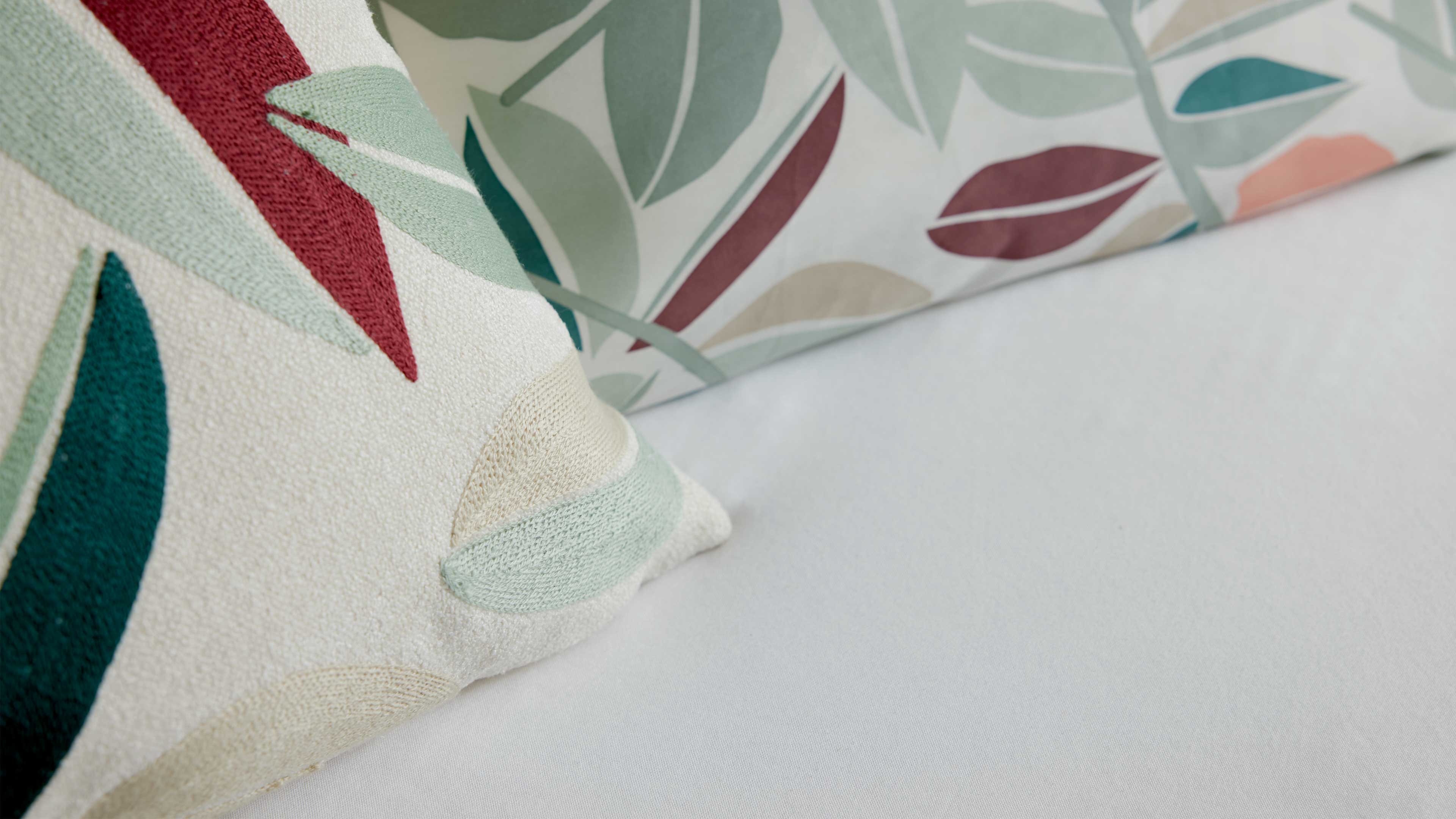 Vordergrundkissen mit applizierten Blattformen in Grün, Petrol und Weinrot liegt auf hellem Bezug, dahinter weiteres Blattkissen.