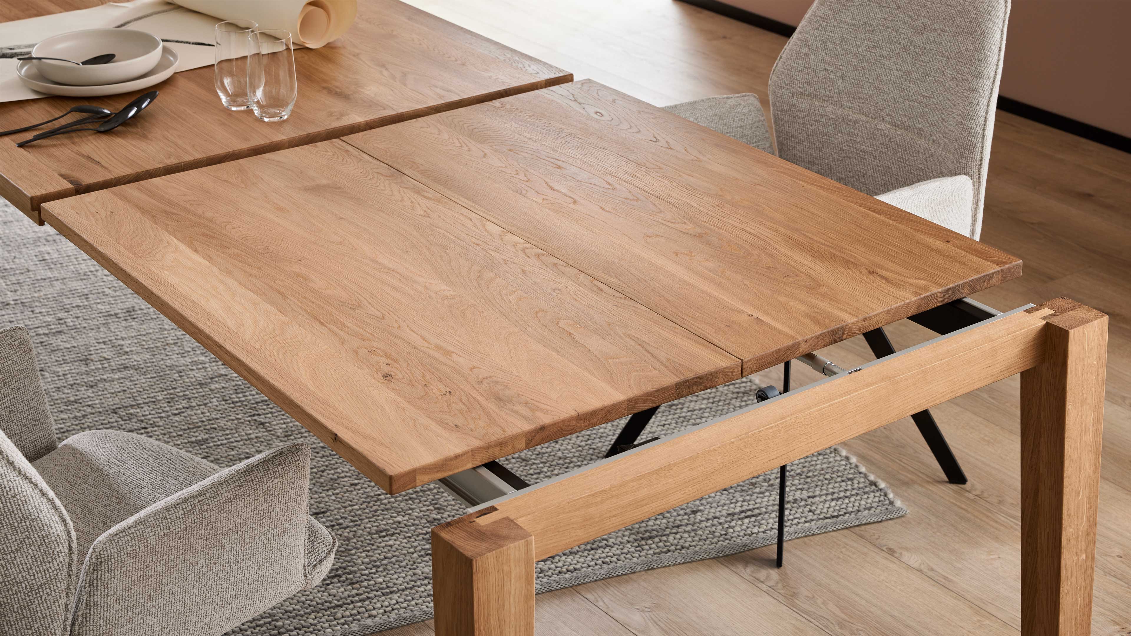 Rechteckiger ausziehbarer Holztisch mit sichtbarer Mittelspalte und Metallauszug, umgeben von hellen, gepolsterten Stühlen.