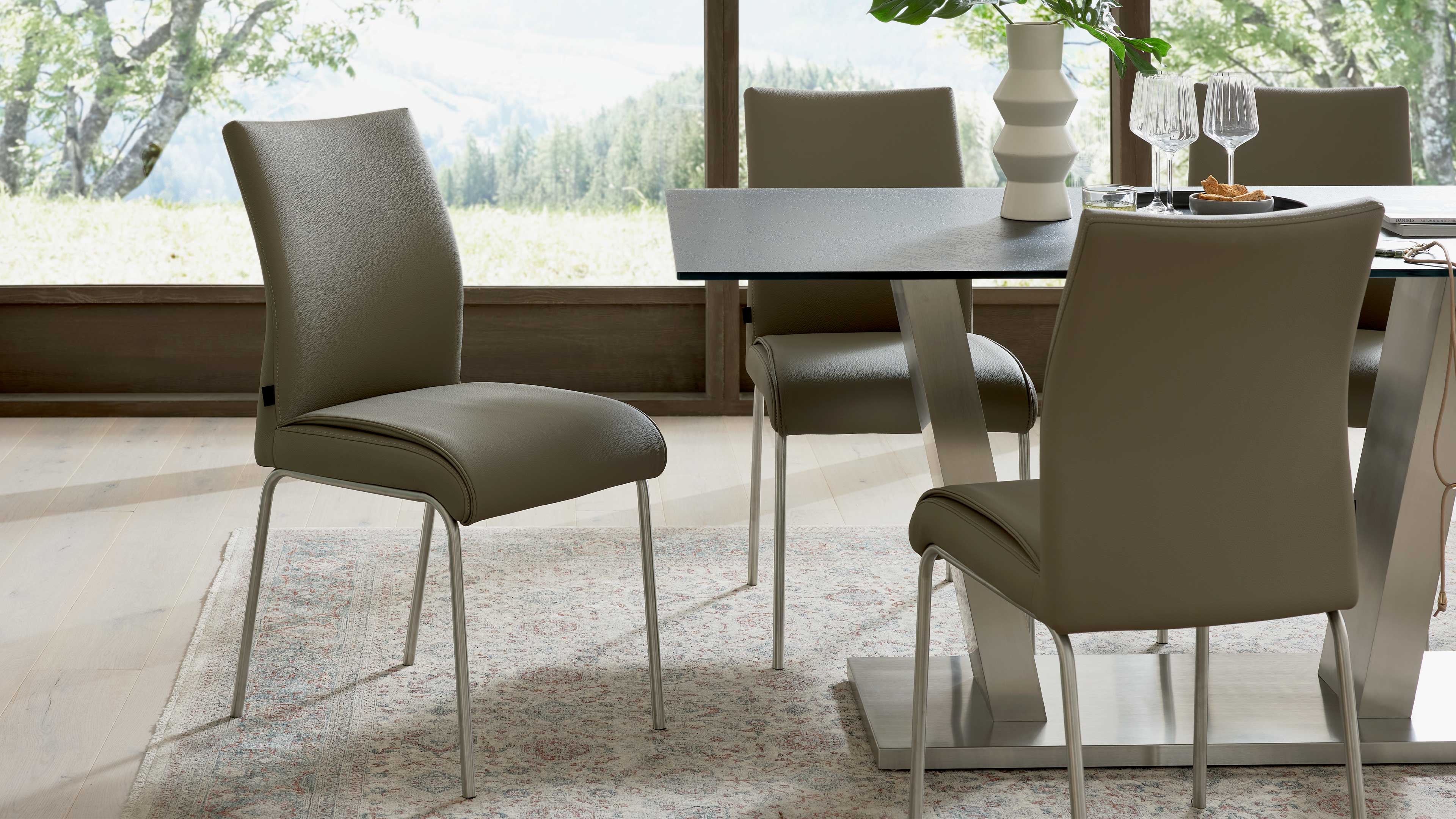 Taupefarbene, gepolsterte Esszimmerstühle mit schlanken Metallbeinen im Vordergrund, dahinter ein Tisch mit Metallfuß, Vase und Gläsern.