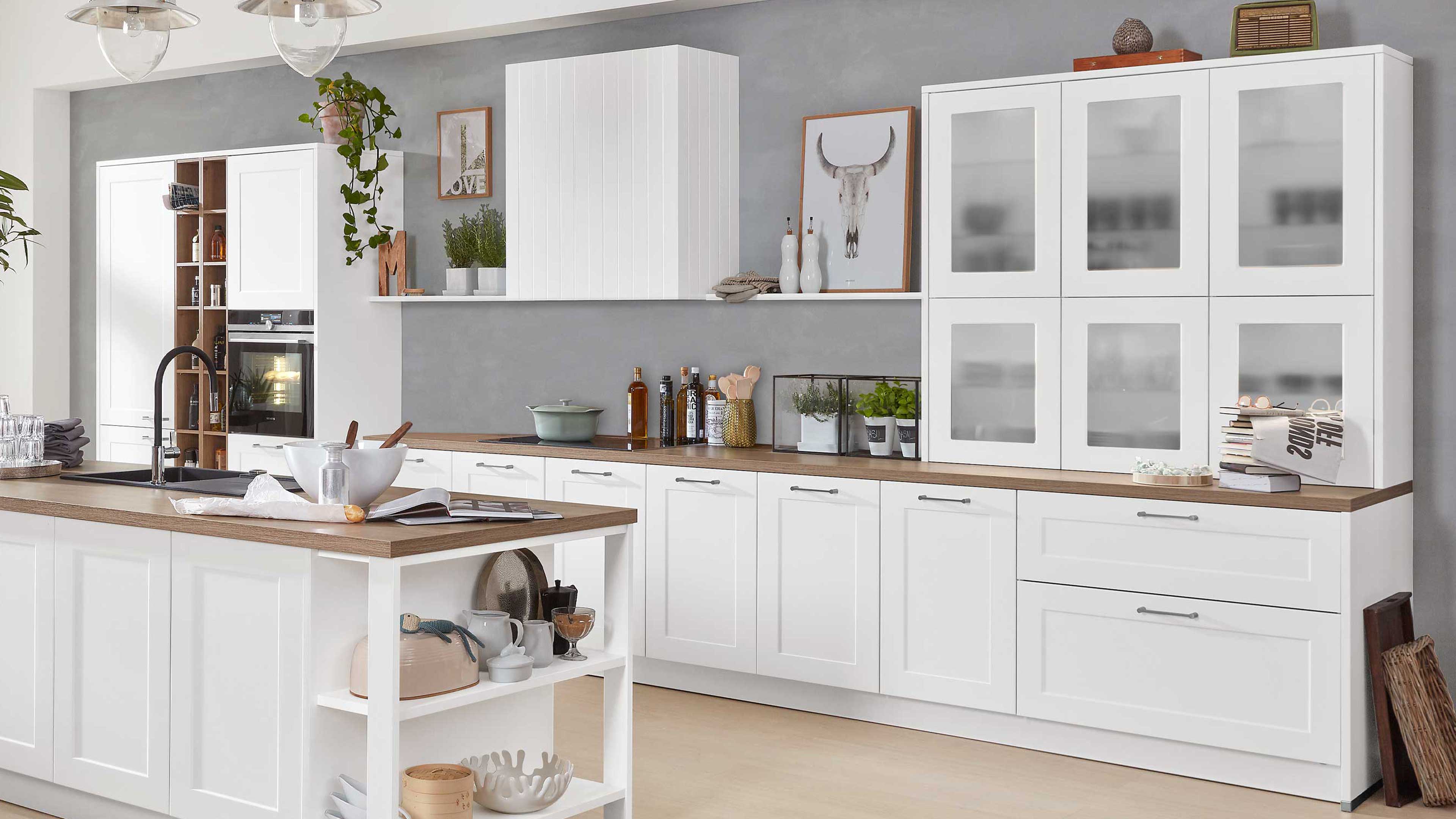 Weiße Küche mit Insel und Holzarbeitsplatten, schwarzer Armatur, Oberschränken mit mattierten Glastüren und dezenter Dekoration.