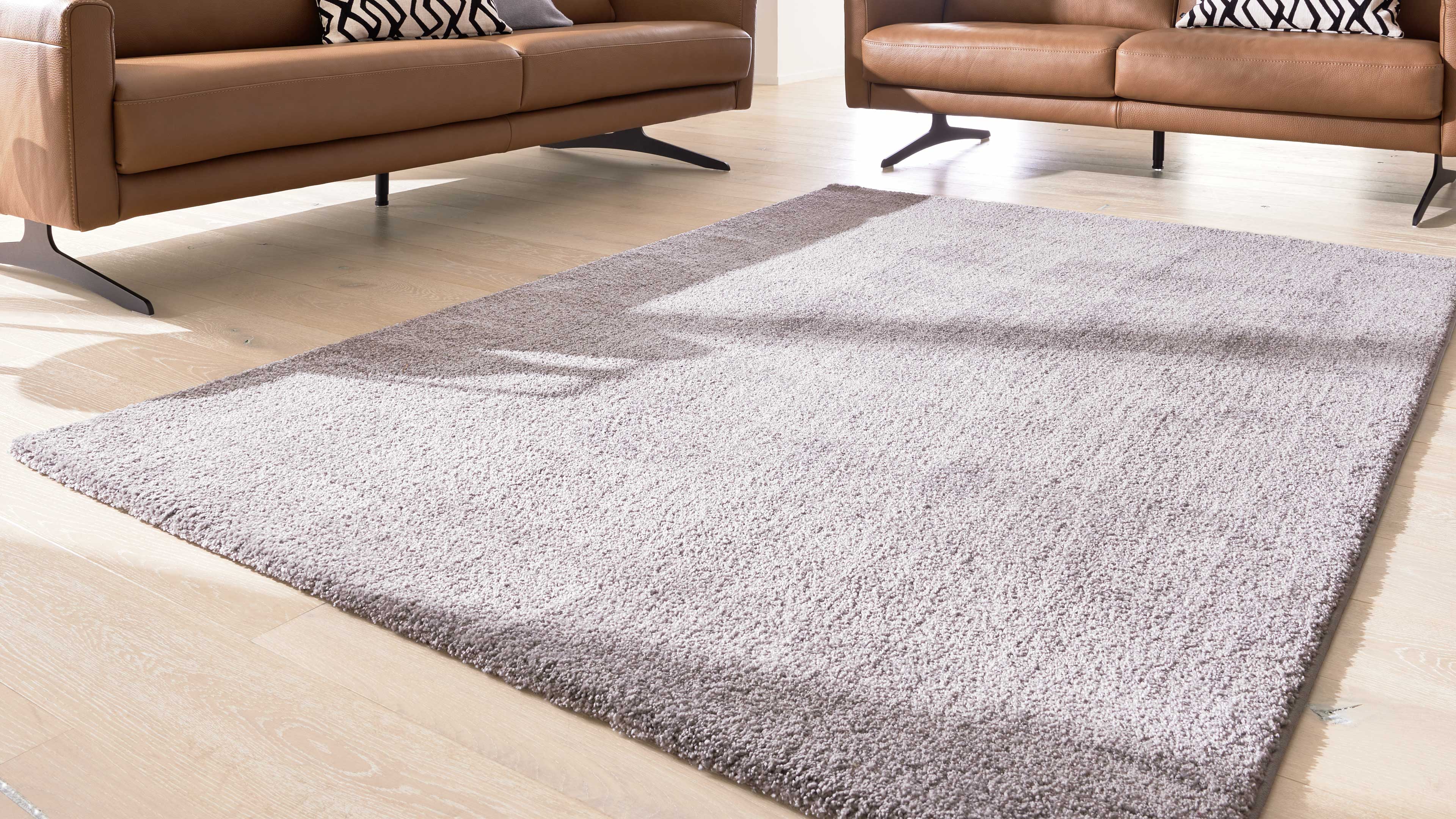 Hellgrauer Hochflorteppich auf hellen Holzdielen, dahinter zwei cognacfarbene Ledersofas mit schwarzen Metallbeinen und gemusterten Kissen.