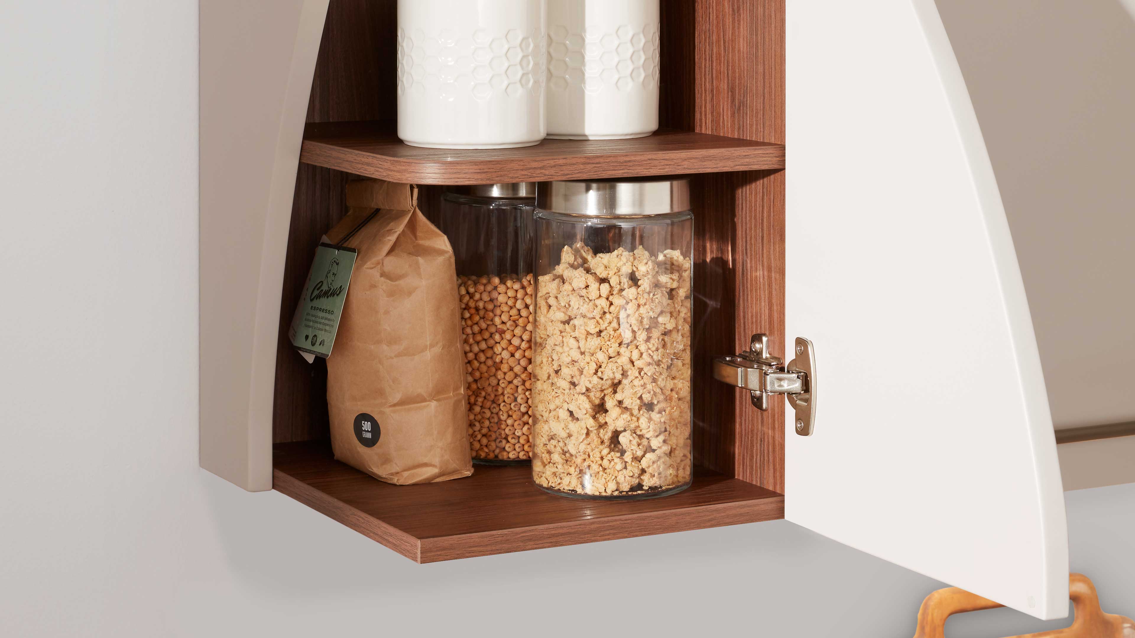 Geöffneter Wandschrank mit weißer Tür, Holzinnenraum, Metallscharnier und zwei Glasbehältern mit Müsli und brauner Papiertüte.