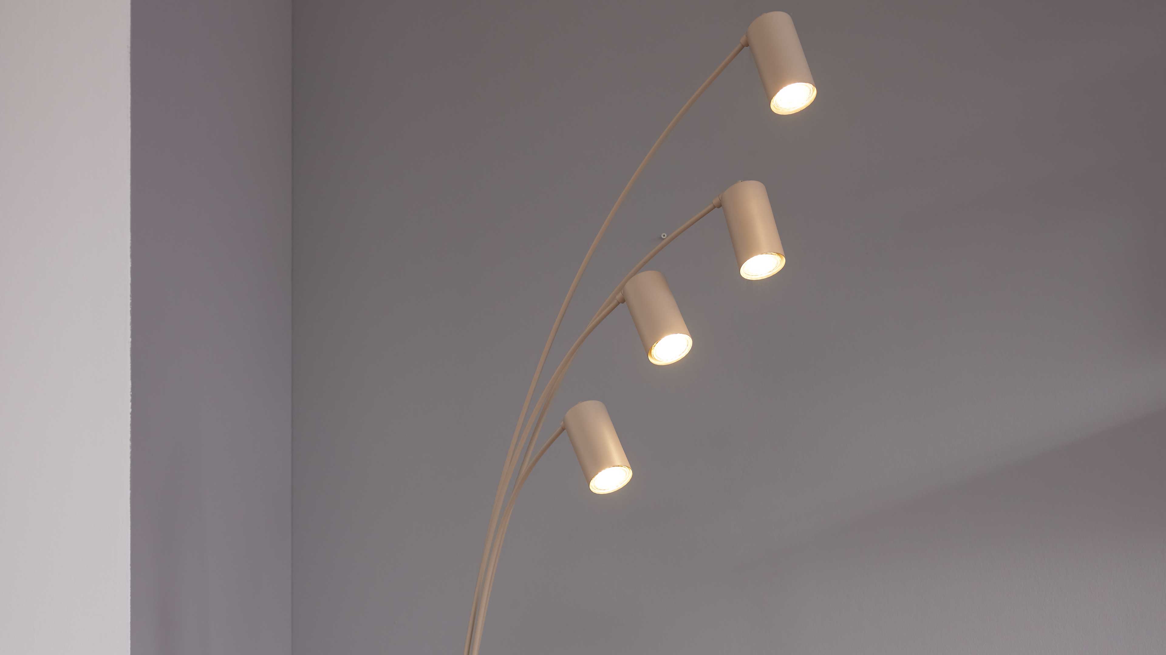 Vier zylindrische, nach oben gebogene Leuchten an gebogenen Armen, eingeschaltet, vor einer hellgrauen Wand und einer weißen Raumkante.