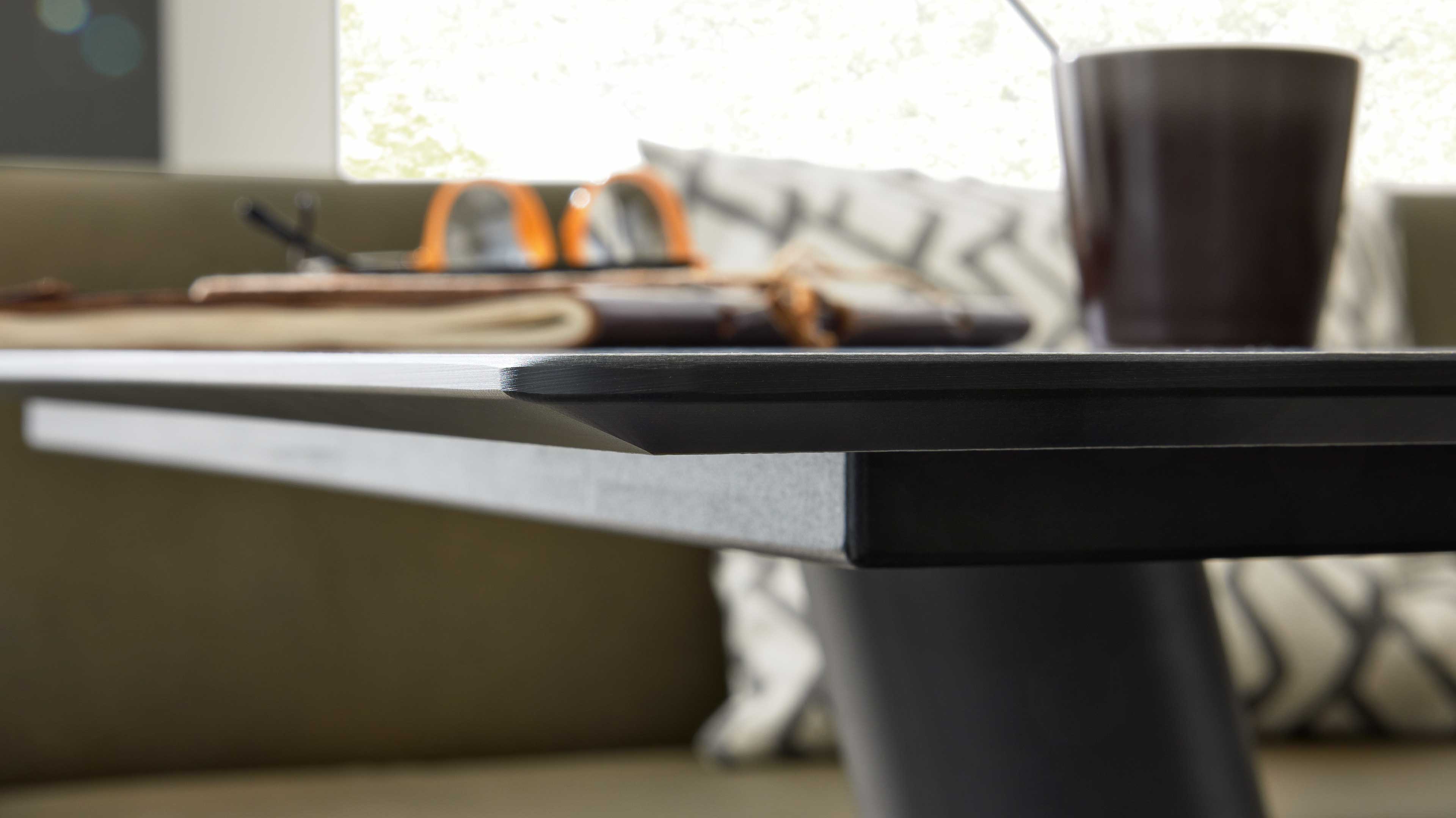 Dunkle rechteckige Tischplatte mit abgeschrägter Kante und rundem Fuß, im Hintergrund unscharf Tasse, Brille, Notizbuch und Kissen.