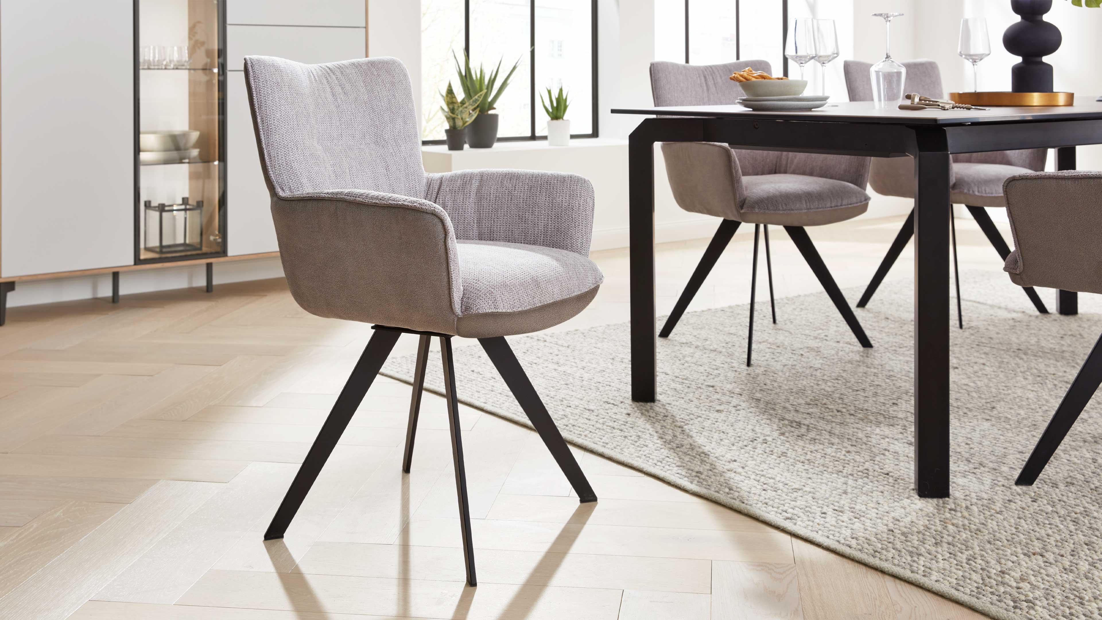 Gepolsterter hellgrauer Armlehnstuhl mit strukturierter Oberfläche und schwarzen Metallbeinen vor einem Esstisch mit Stühlen.