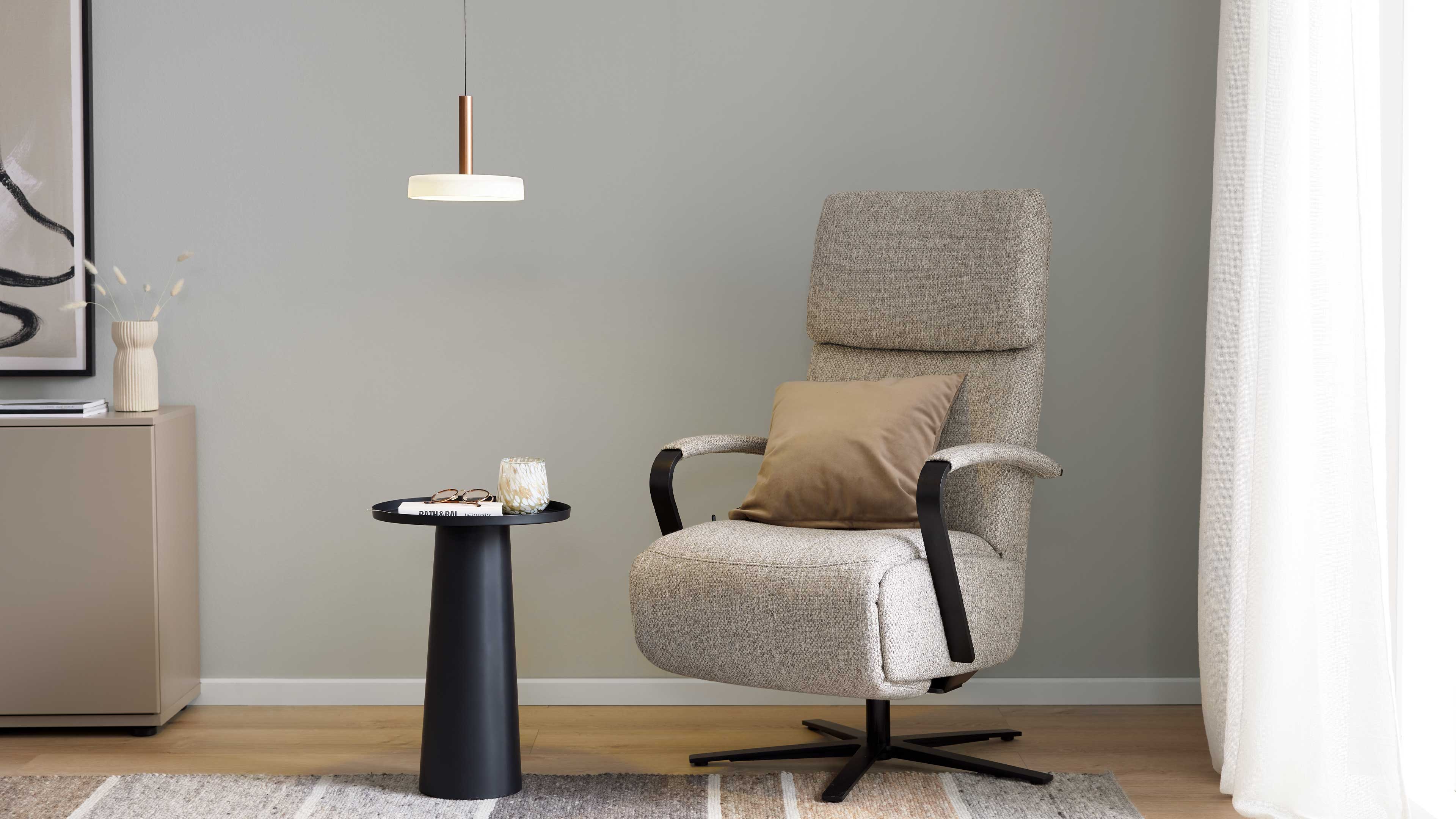 Hellgrauer, gepolsterter Sessel mit braunem Kissen und schwarzen Armlehnen, daneben schwarzer Beistelltisch mit Buch und Kerze.